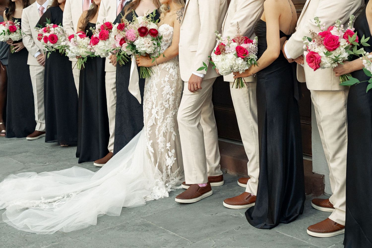 Wedding party in navy gowns and tan suits holding red and white bouquets lined up for formal photos.
