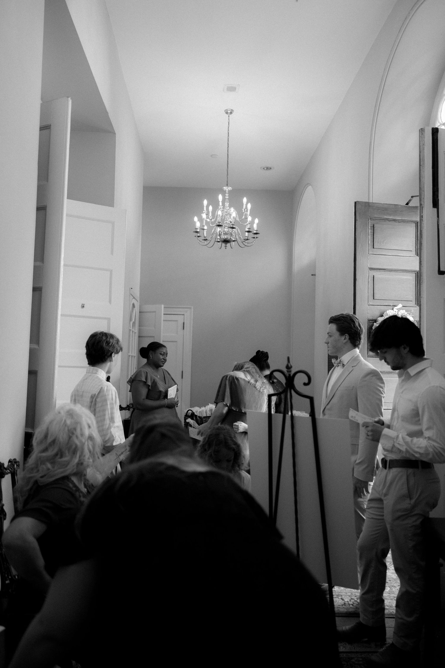 Black and white photo of wedding guests gathering in a chandelier-lit hallway before the ceremony begins.