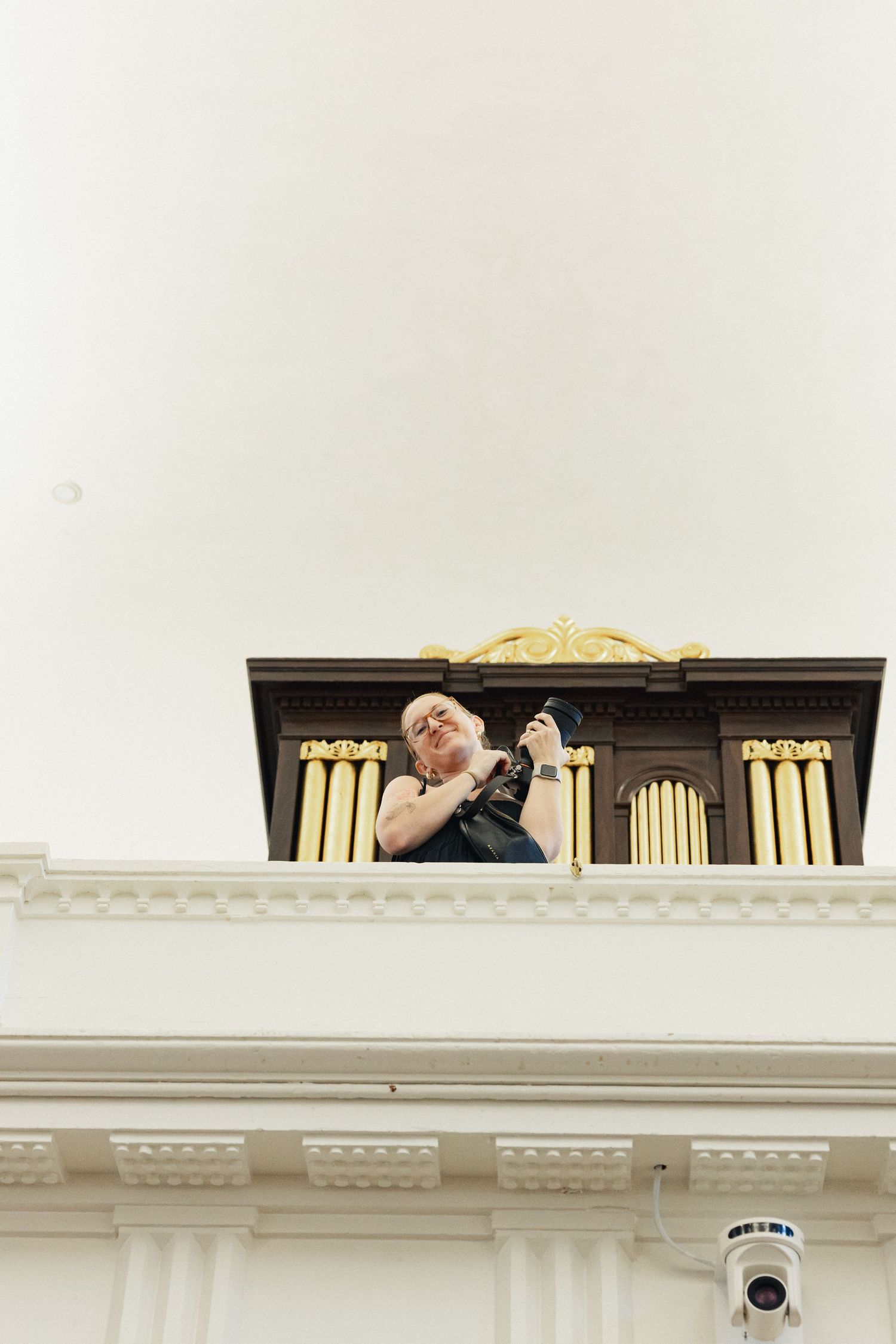 Woman photographing from a church balcony near a ornate wooden organ with gold pipes.