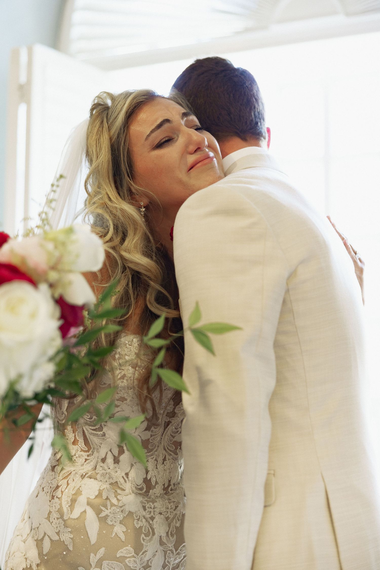 Bride with wavy blonde hair embraces groom in ivory suit, holding red and white bouquet at wedding ceremony.