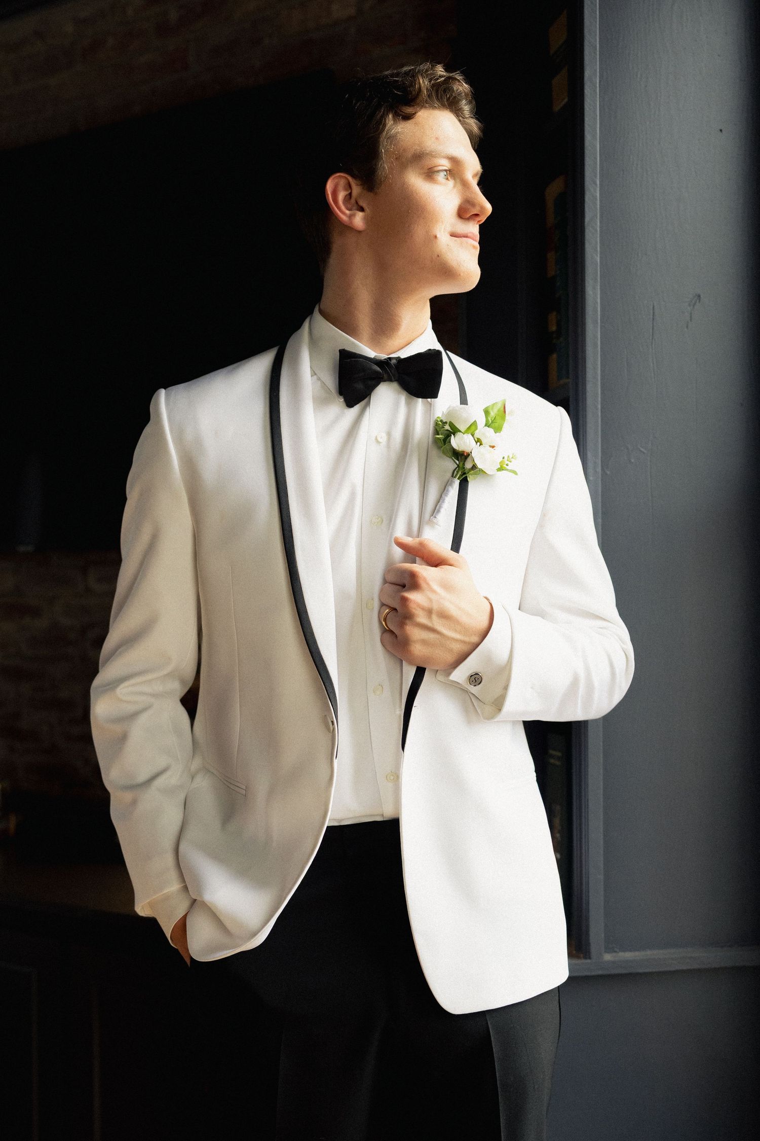 Groom in white tuxedo jacket with black bow tie and floral boutonniere posing elegantly indoors.