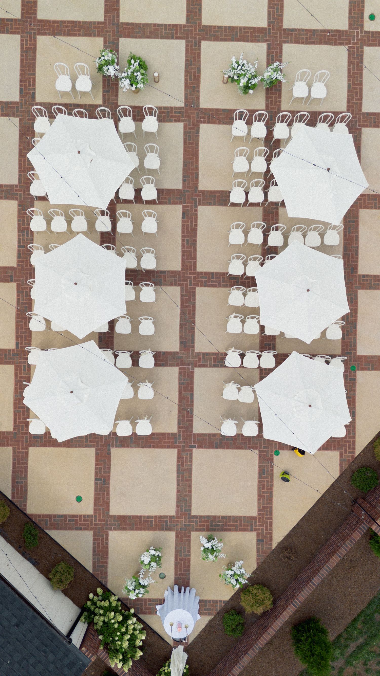Aerial view of an elegant outdoor event setup with white patio umbrellas, chairs, and floral arrangements on a brick plaza.