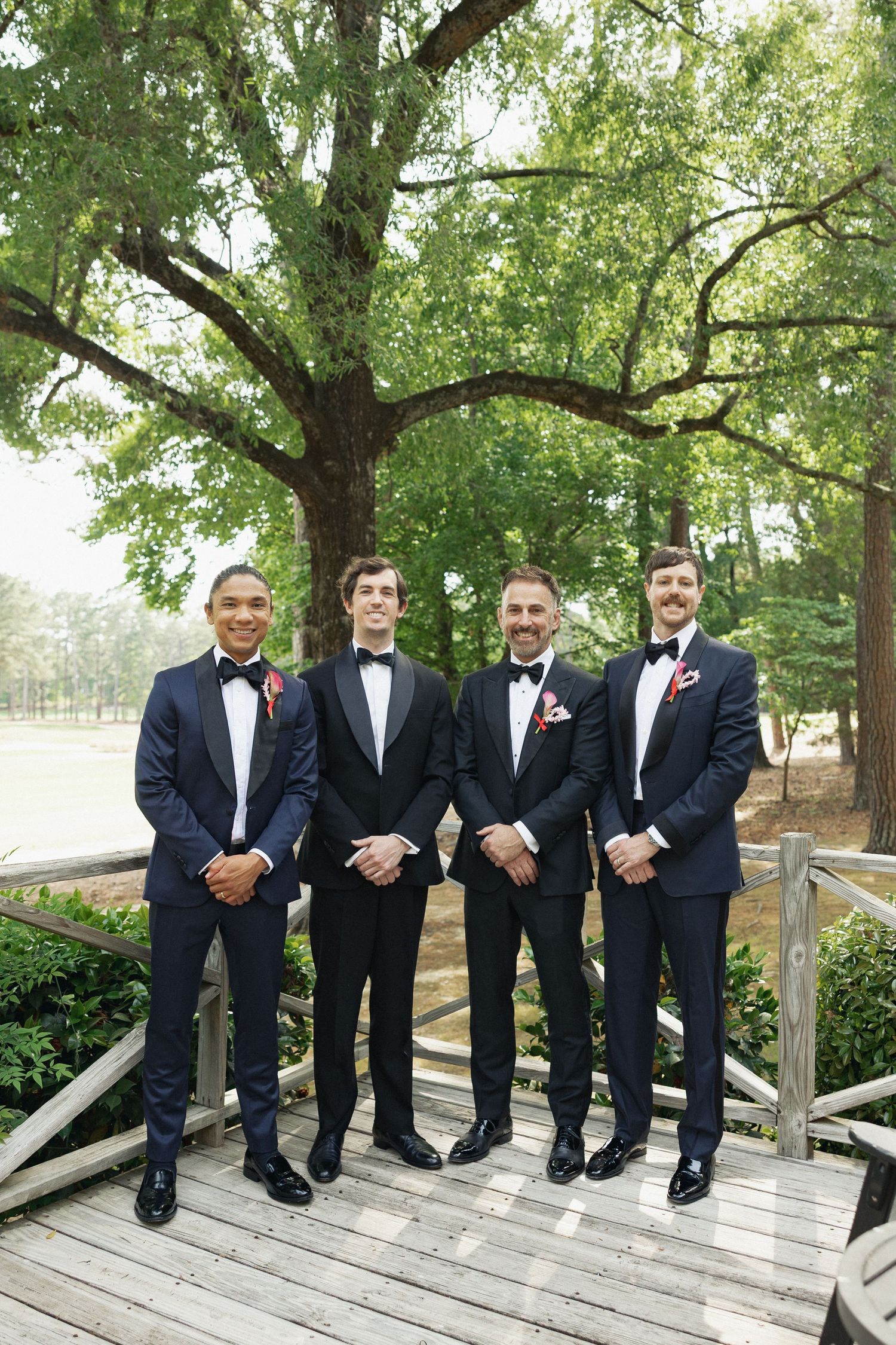 Four groomsmen in navy tuxedos with red boutonnieres pose on a wooden bridge surrounded by lush green trees.
