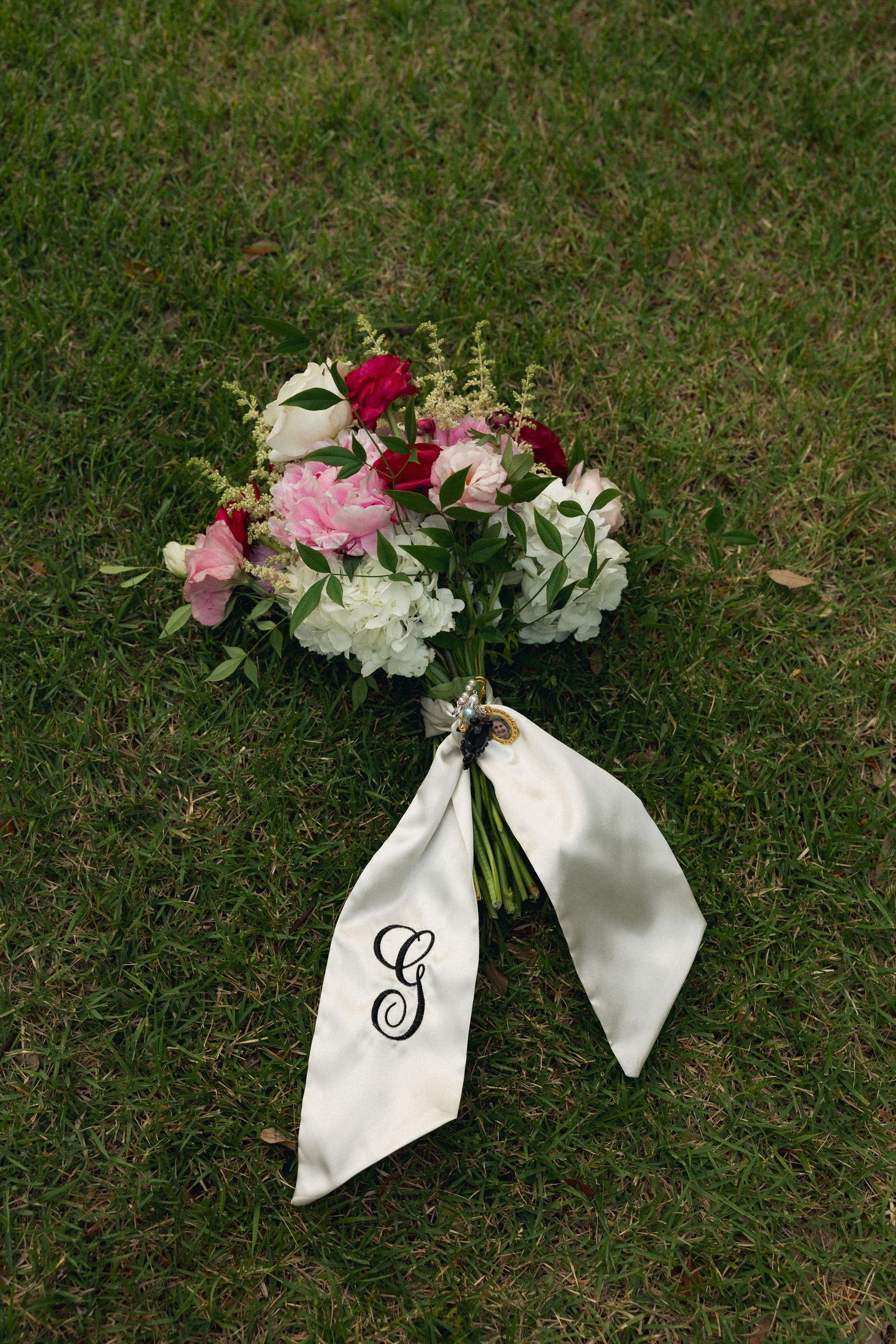 Bridal bouquet of pink, red, and white flowers with monogram G satin ribbon on green grass.