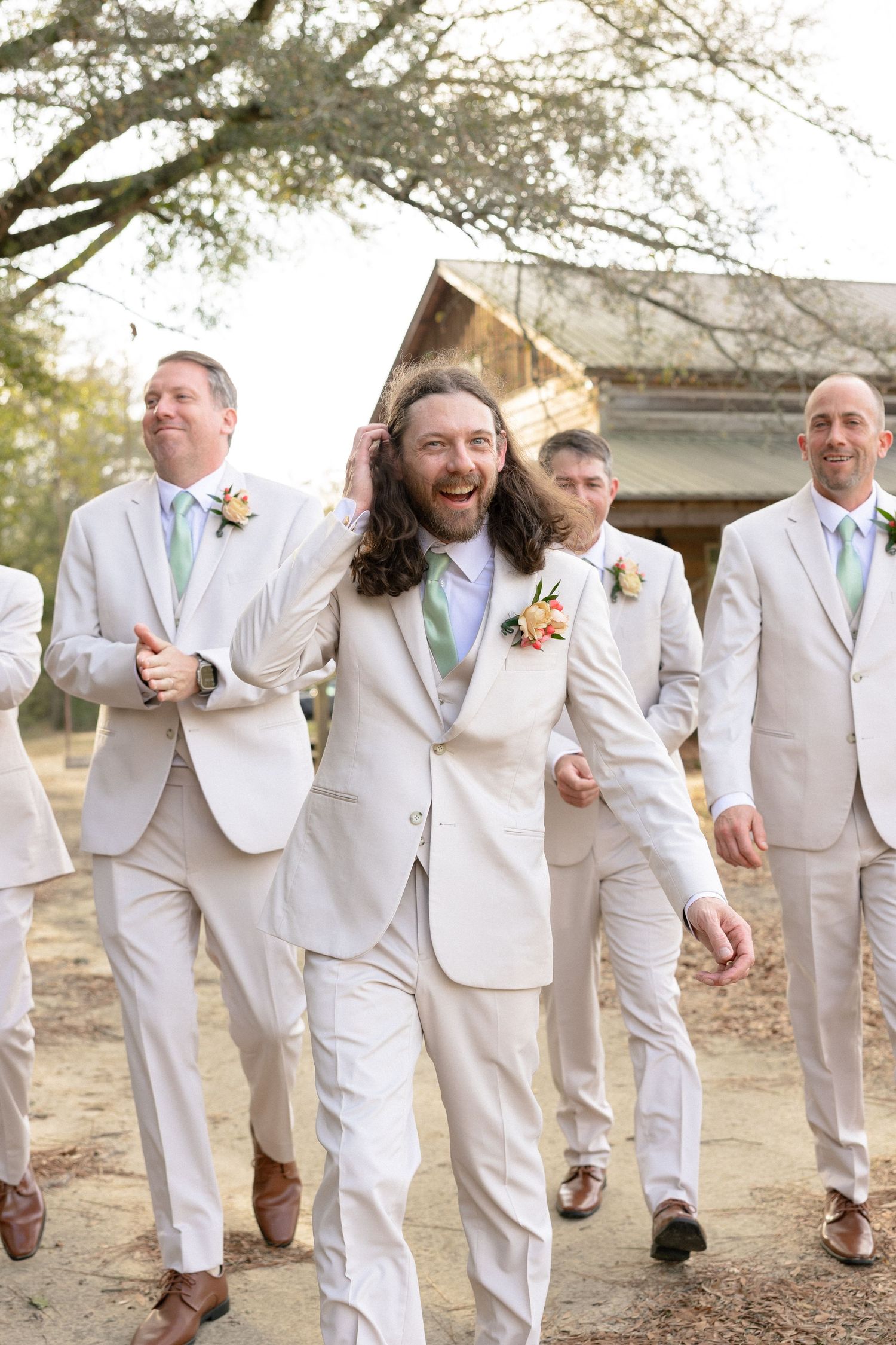 Groomsmen in cream suits laugh and walk together outside a rustic barn wedding venue on a sunny day.