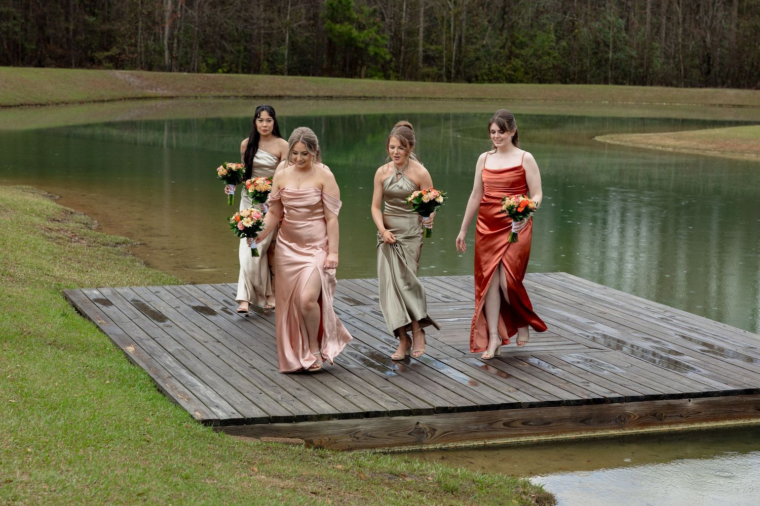 Four bridesmaids in rust, blush, and gold satin dresses walk across a wooden dock beside a calm green lake.