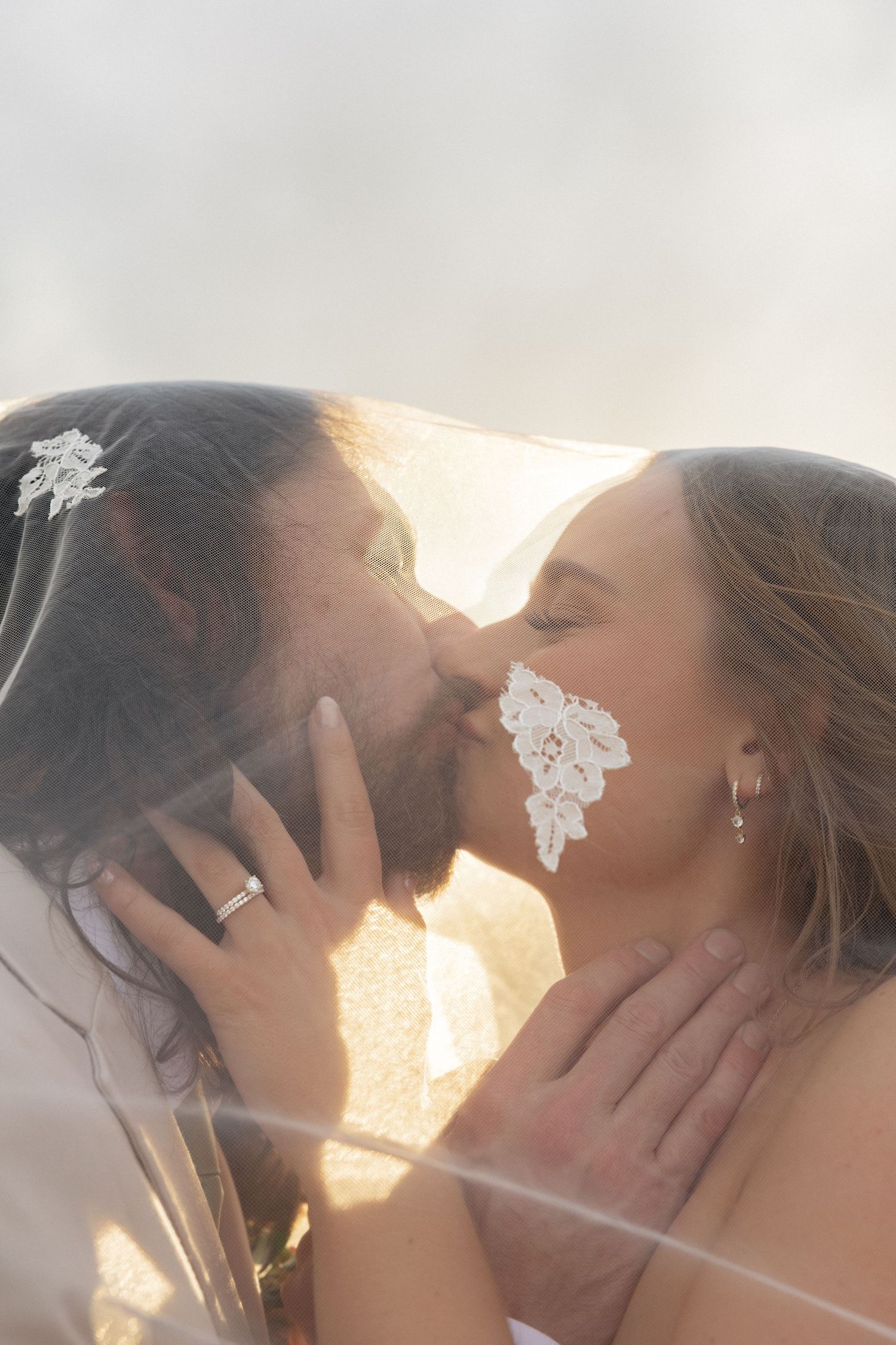 Bride and groom kiss under lace veil with warm backlight, wedding rings visible.