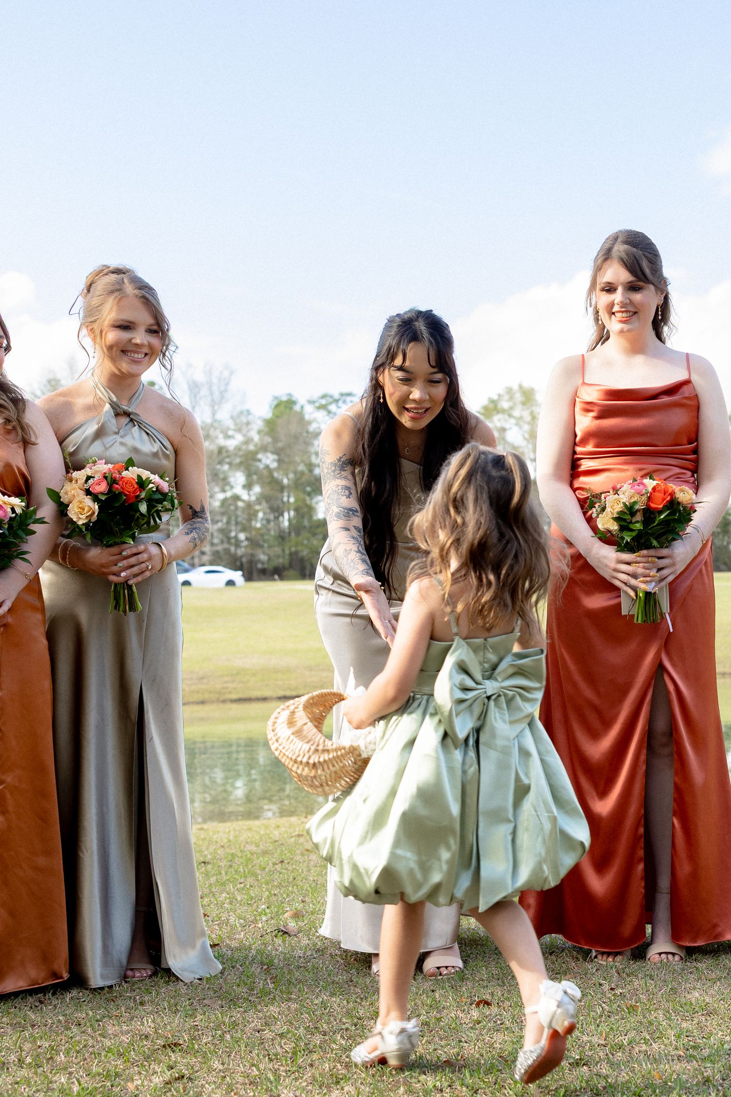 A flower girl in mint dress with bow interacts with bridesmaids wearing rust and champagne satin gowns at outdoor wedding.