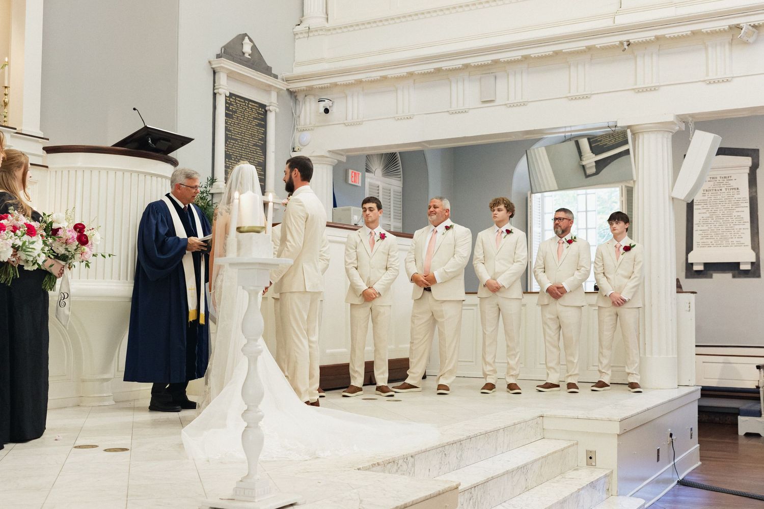 Groom in cream suit stands at altar with groomsmen as officiant in blue robe leads the ceremony.