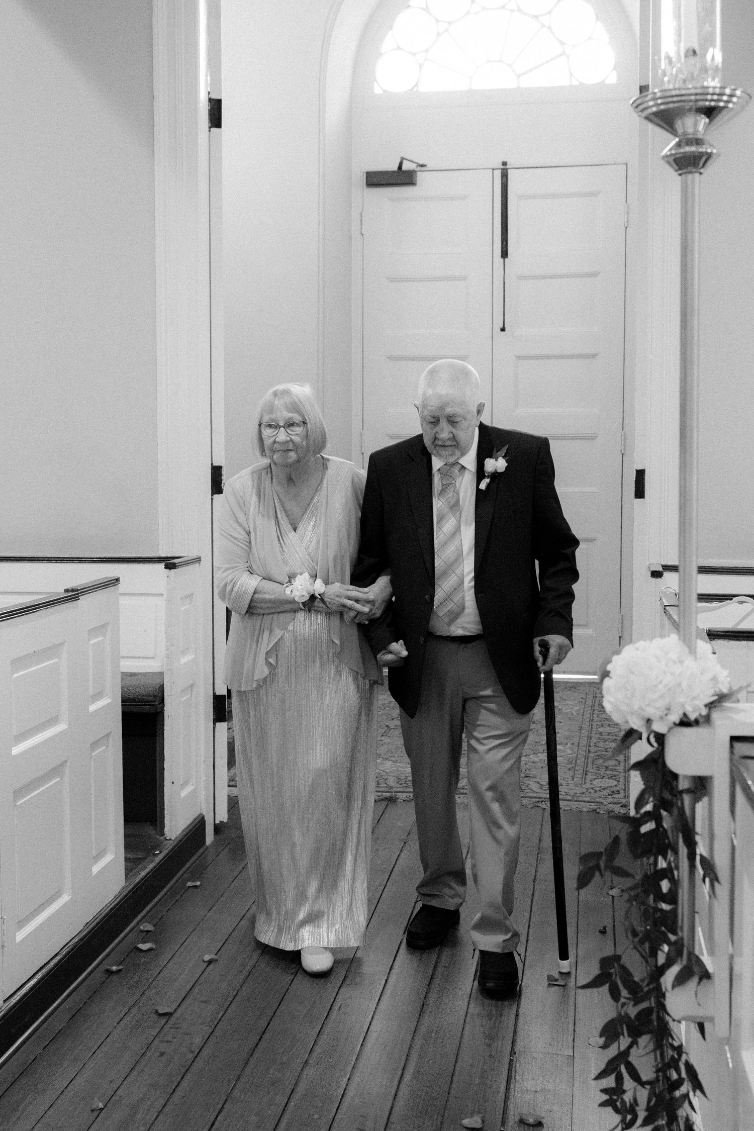 Elderly couple walking down church aisle together, man using cane, woman holding flowers, black and white wedding photo.