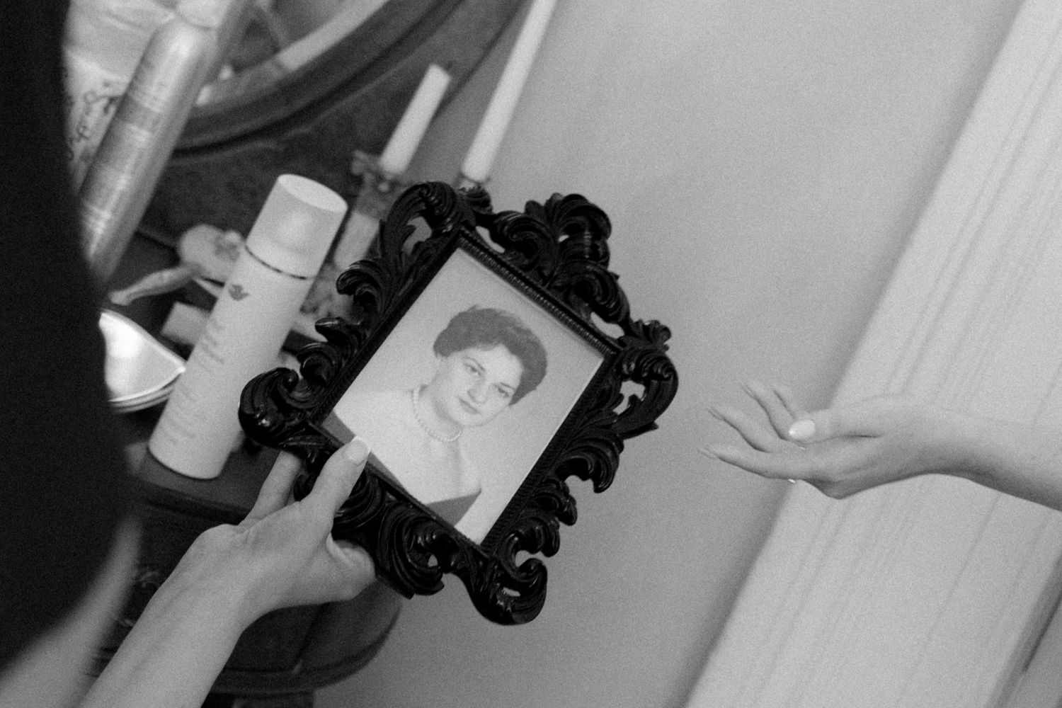 Black and white photo of a person holding an ornate framed portrait near a mirror with candles in background.