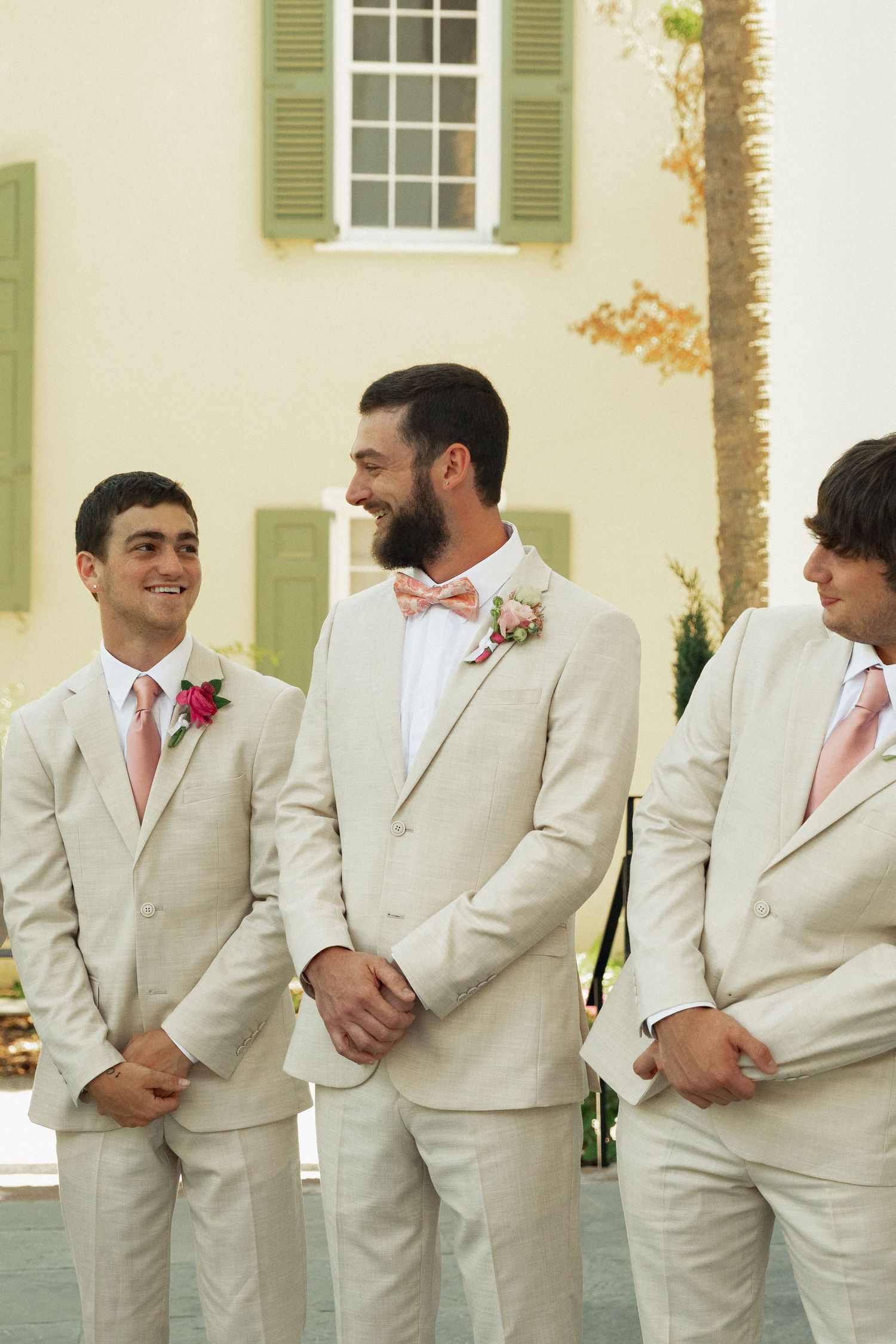 Groom and groomsmen in tan suits with pink bow ties and boutonnieres laughing together outdoors.