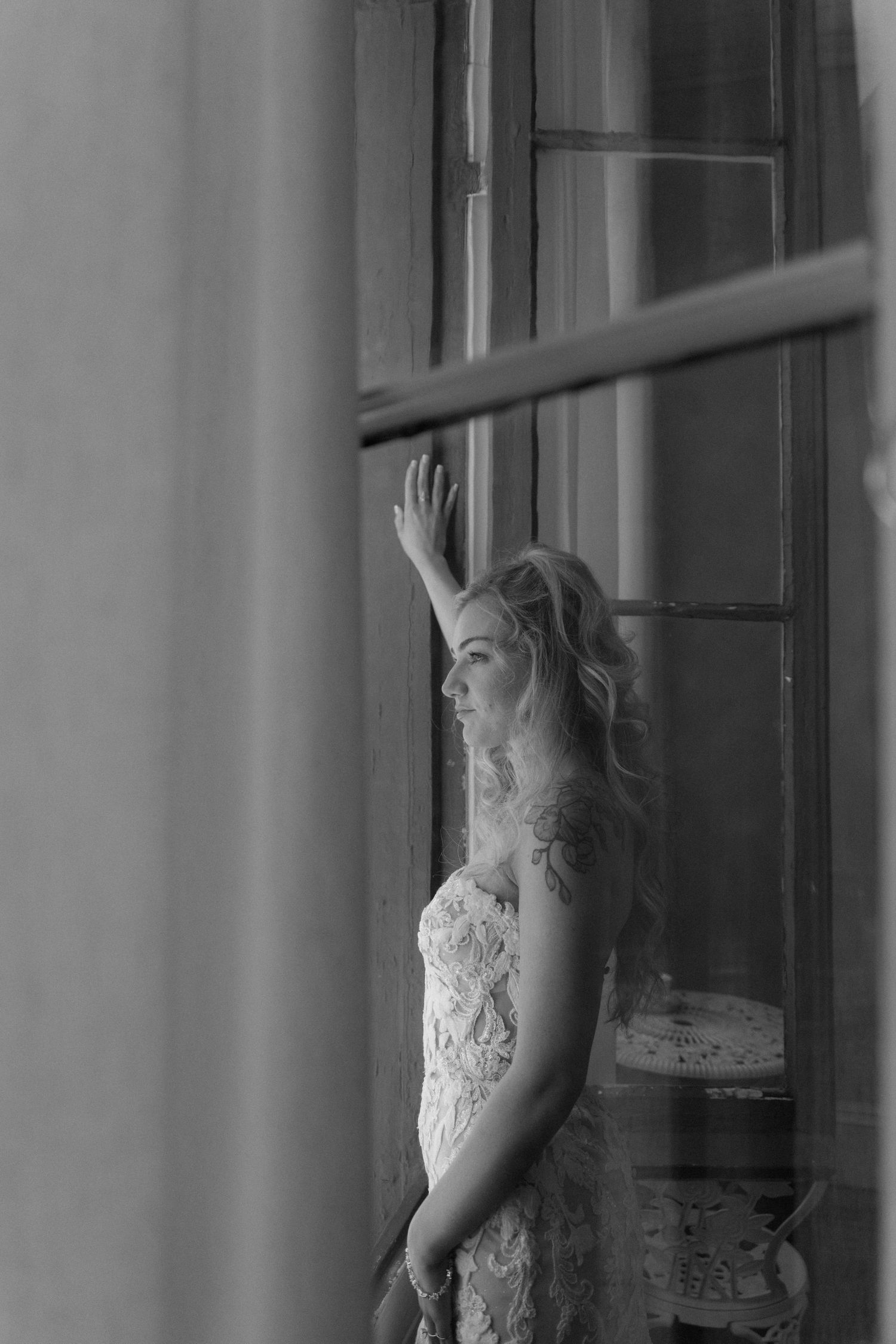 Black and white photo of blonde bride in lace gown touching vintage window in elegant building.