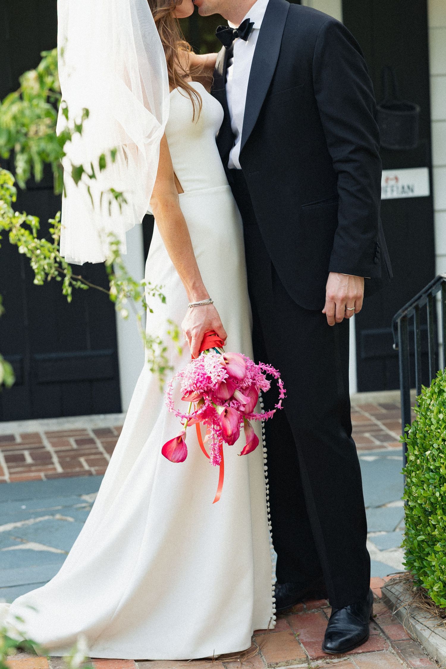 Bride and groom kiss outdoors, bride holding vibrant pink floral bouquet, both dressed in classic white and black wedding attire.