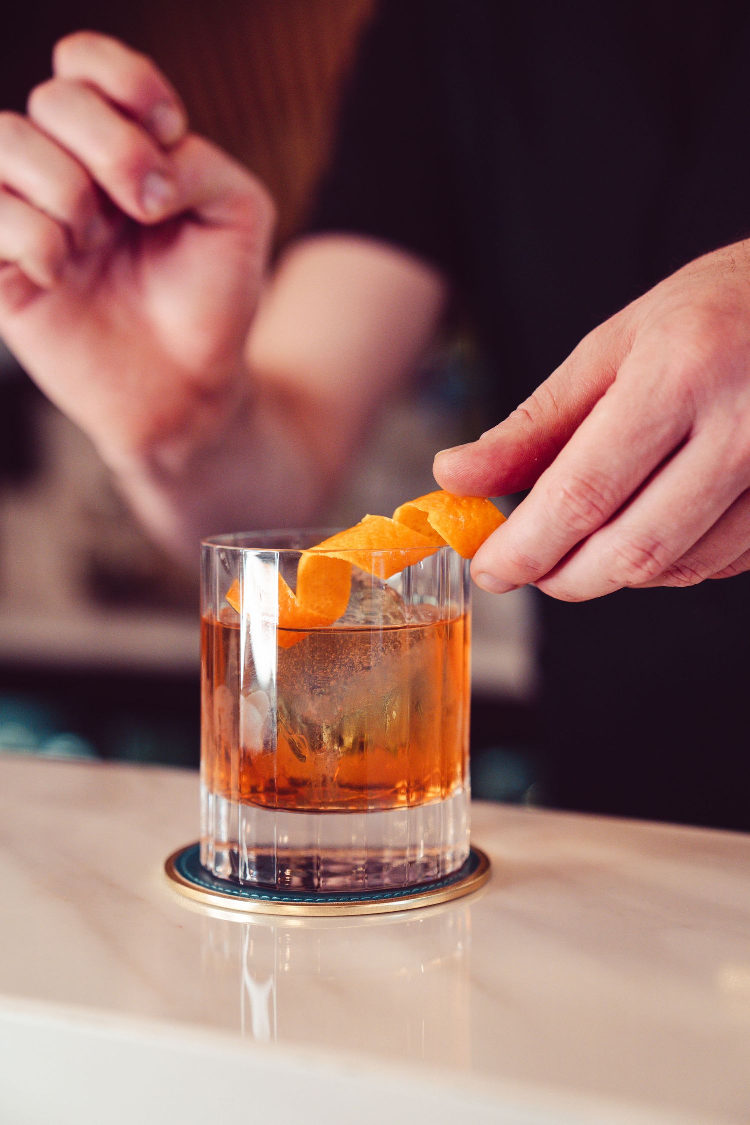 Hands garnishing an old fashioned cocktail with orange peel in a crystal glass on white marble surface.