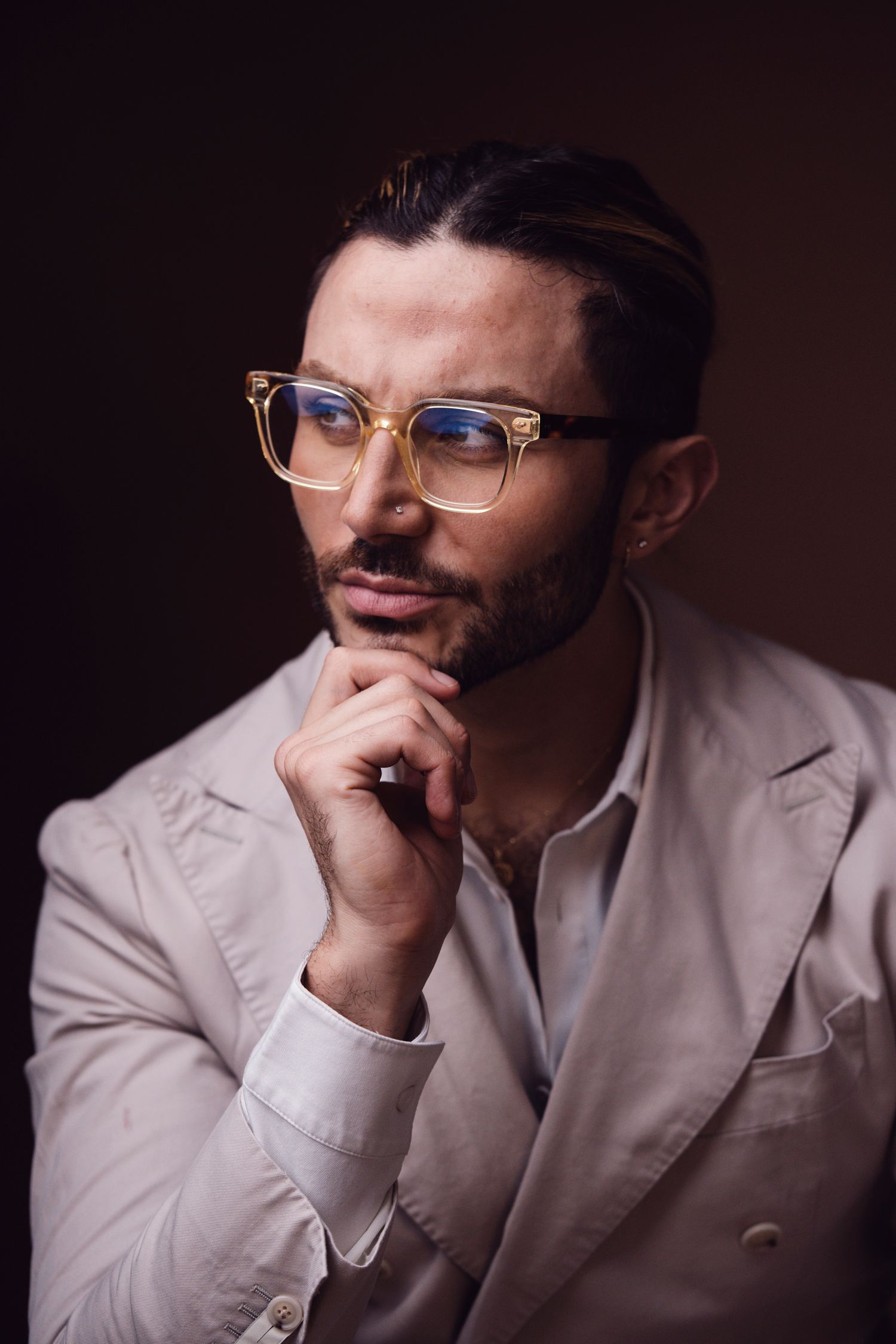 Thoughtful man with glasses and beard in white blazer posing with hand on chin against dark background.