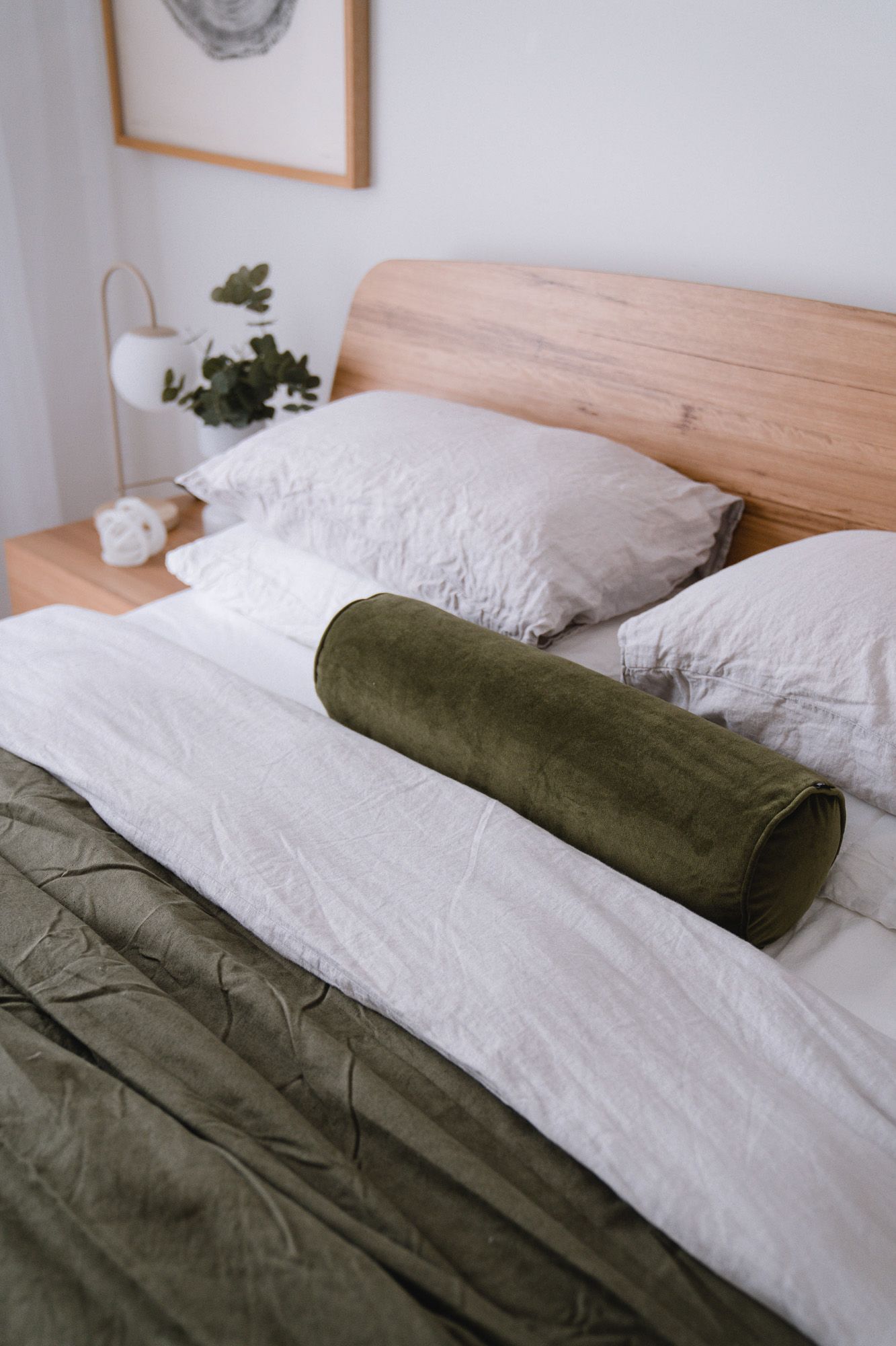 Cozy bedroom with wooden headboard, white linen pillows, green velvet bolster pillow, and olive duvet on a neatly made bed.