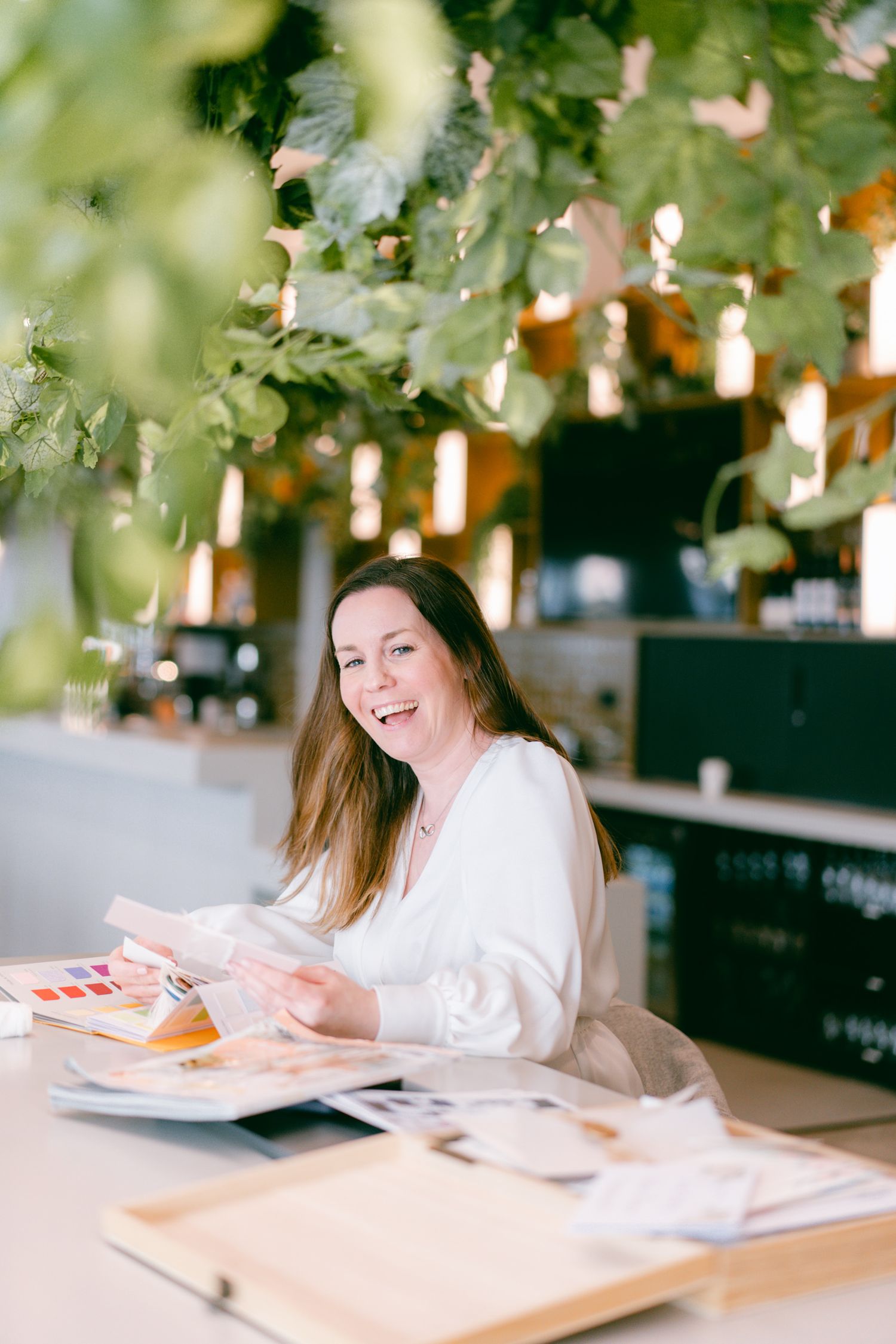 Woman smiling at a cafe table, reviewing colorful design swatches surrounded by lush green plants.