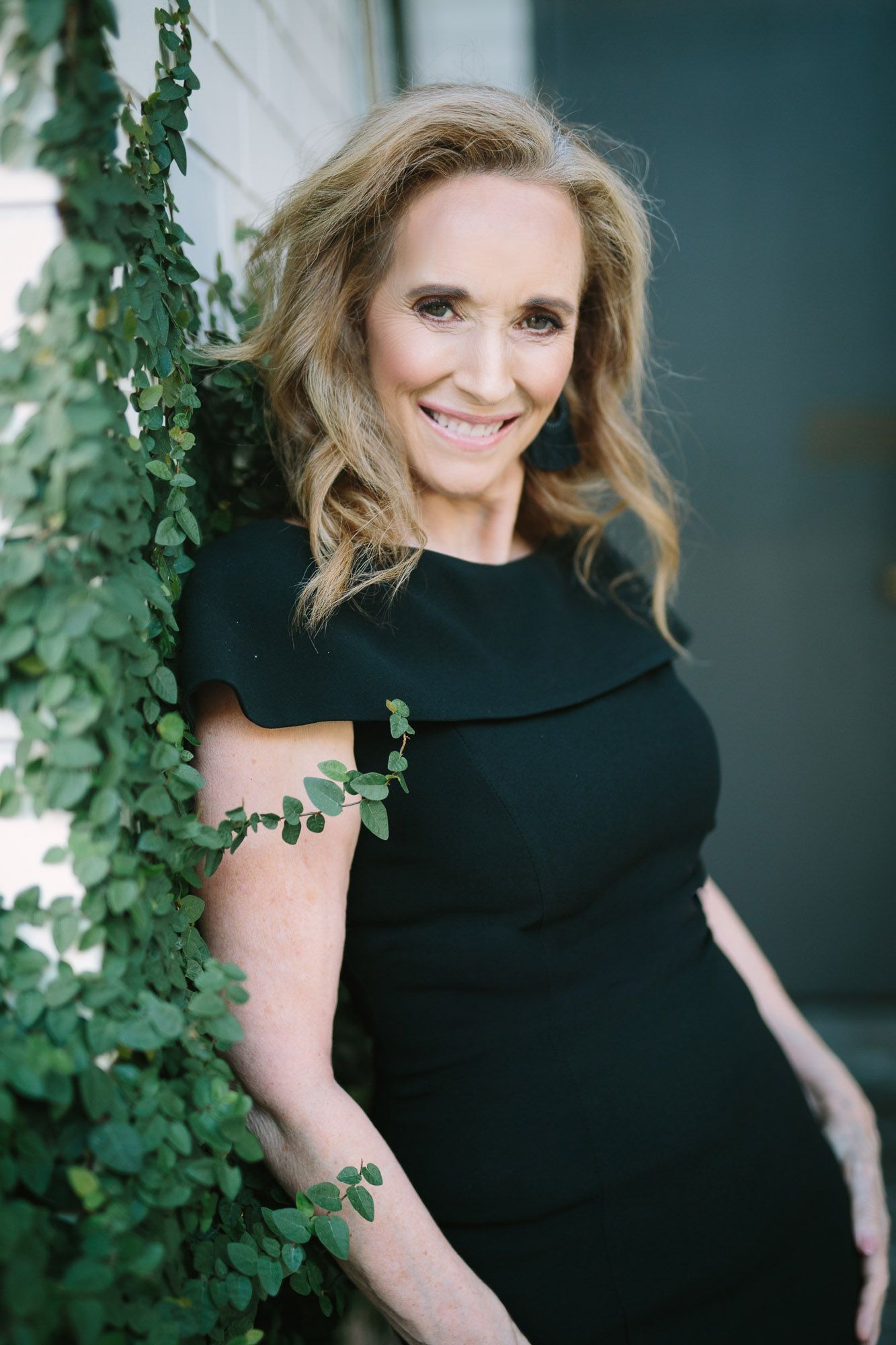 A woman in a black dress poses against a wall with climbing green vines in an elegant outdoor portrait.