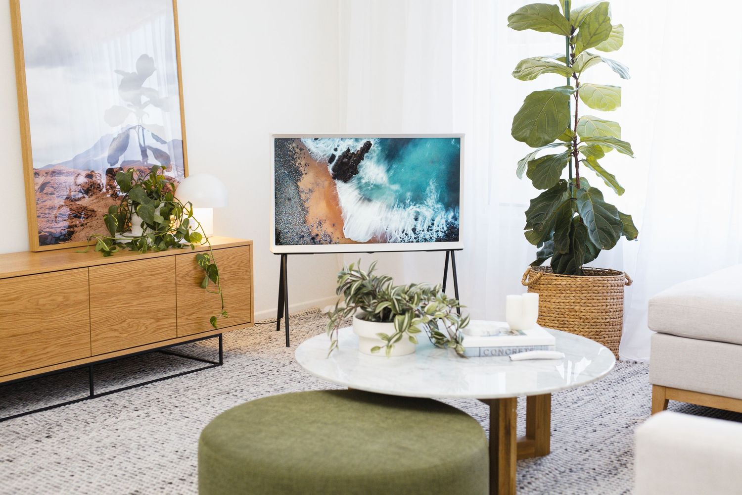 Modern living room with Samsung The Frame TV on easel stand, wood sideboard, fiddle leaf fig, and green ottoman on neutral rug.