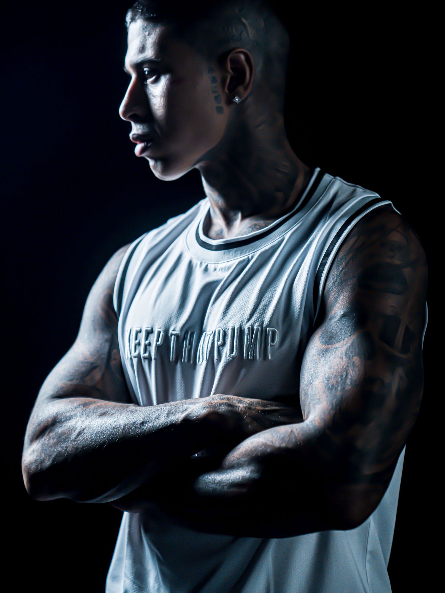 Athletic figure in white tank top showing tattooed arms against dark background.