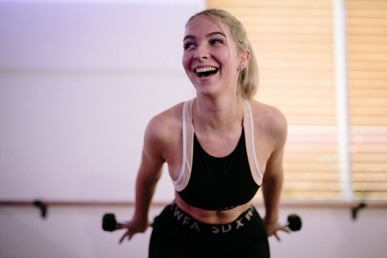 Smiling blonde woman in black sports bra and leggings laughing during a workout in a fitness studio.