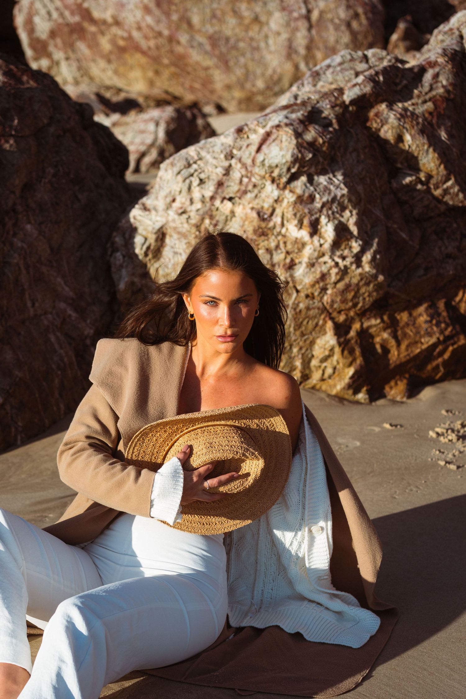 Fashion portrait on beach with tan blazer and white outfit against rocky backdrop.