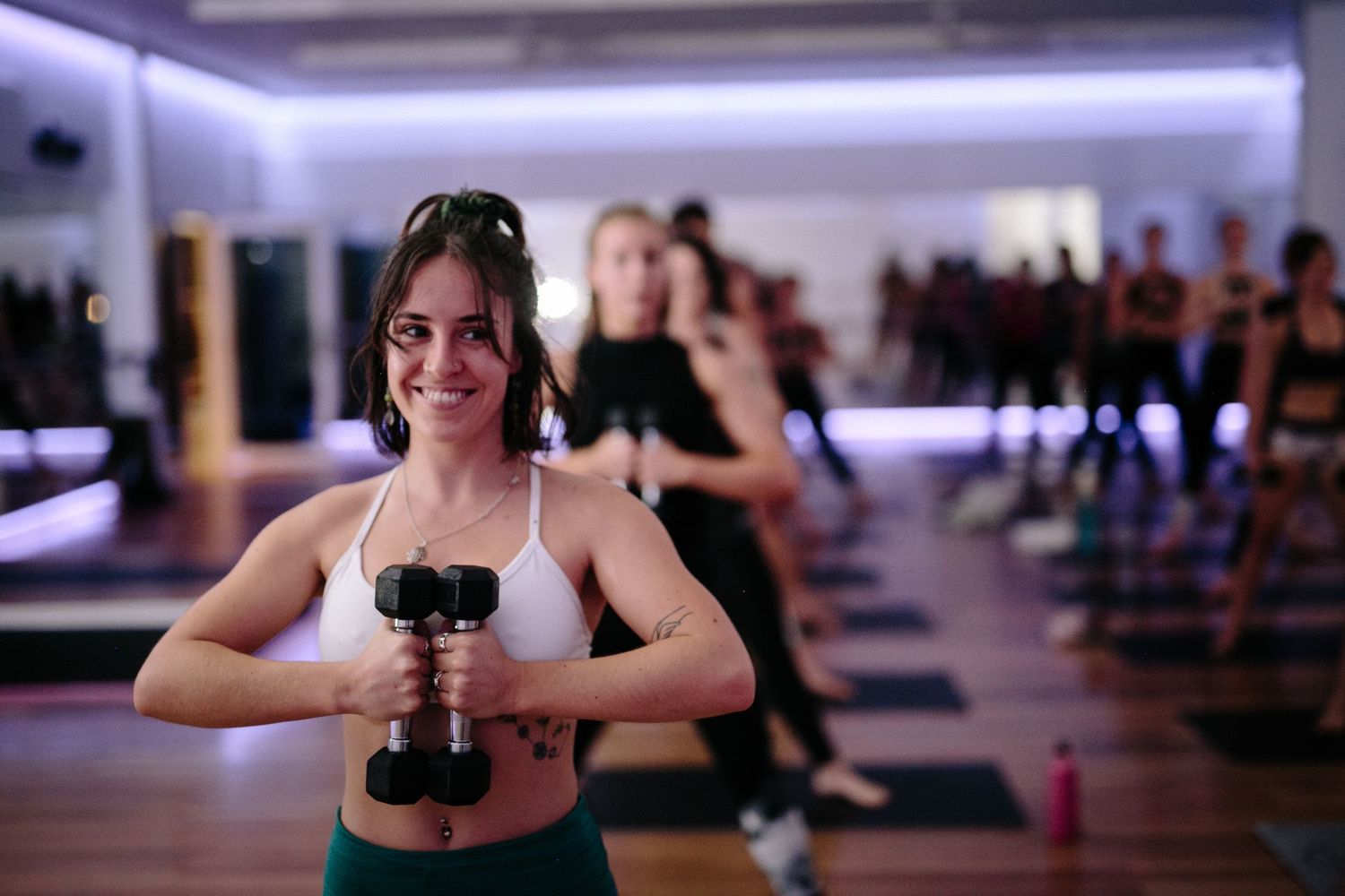 Woman smiling while holding dumbbells in a group fitness class at a modern gym studio.