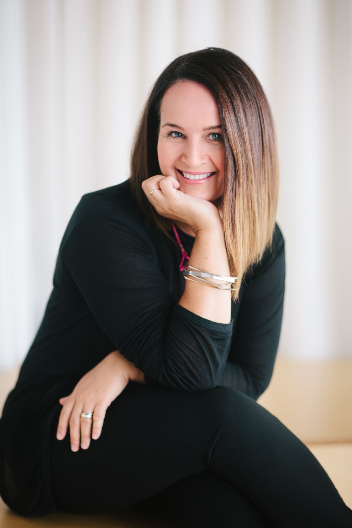 Smiling woman with ombre hair wearing black outfit and bracelets, posing casually against white curtain backdrop.