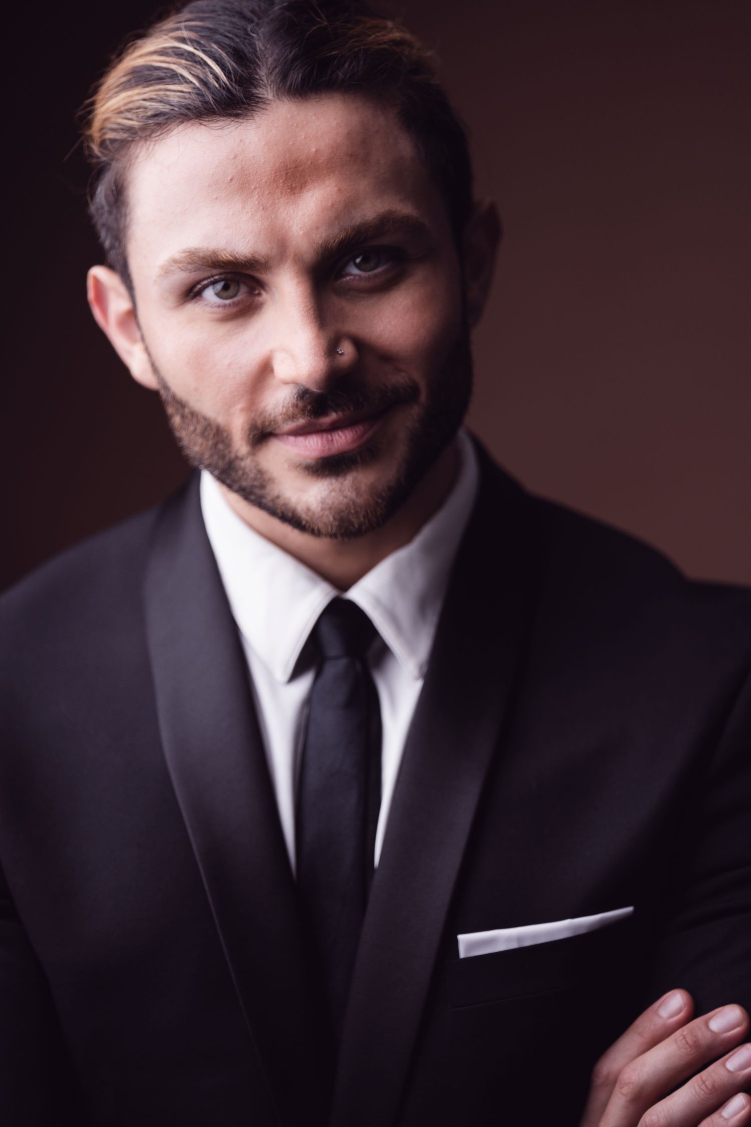 A confident man in a black tuxedo with white pocket square and tie poses against a dark brown background.