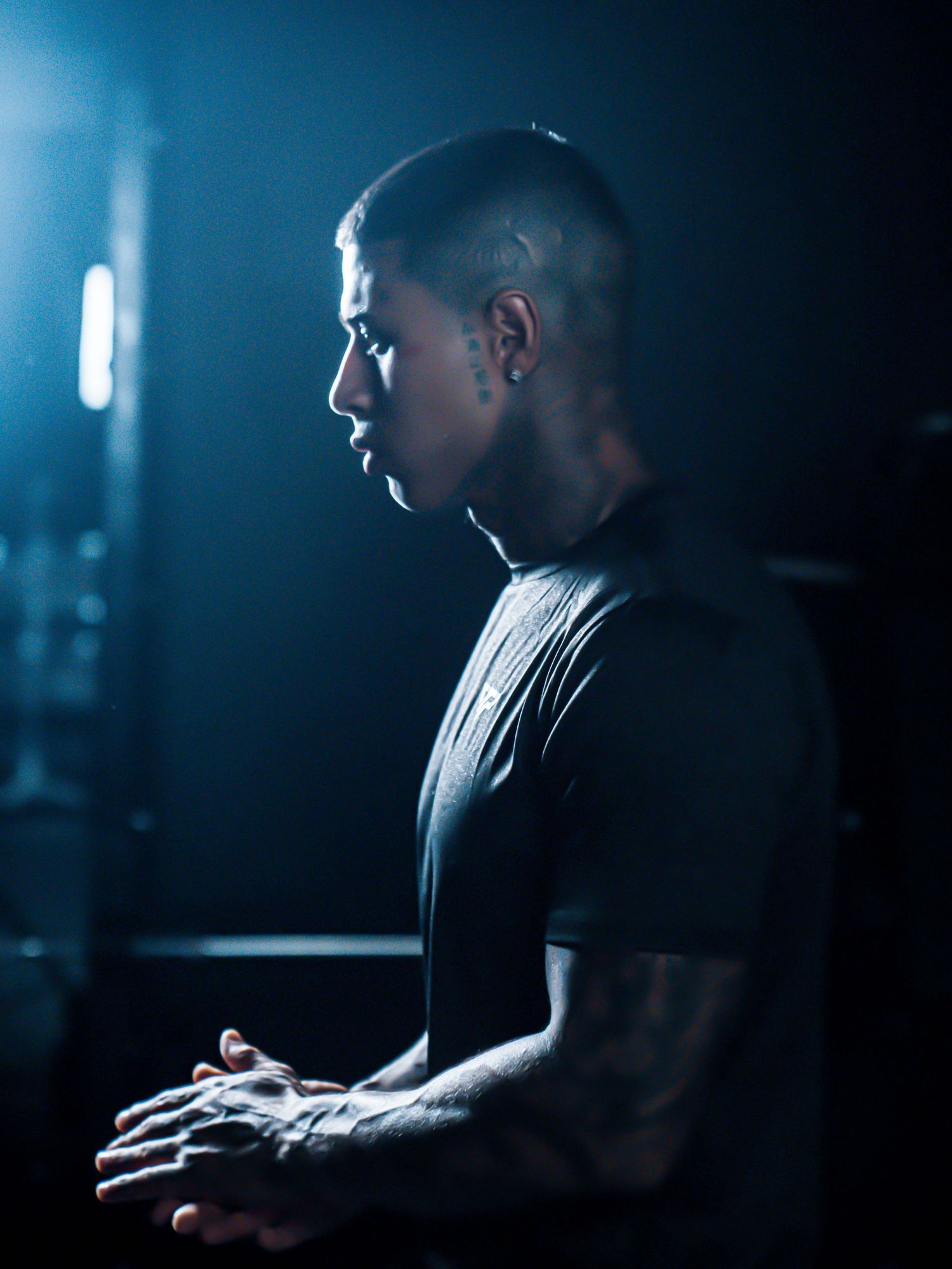 Tattooed man in black shirt with shaved head stands in profile against dark blue moody background.