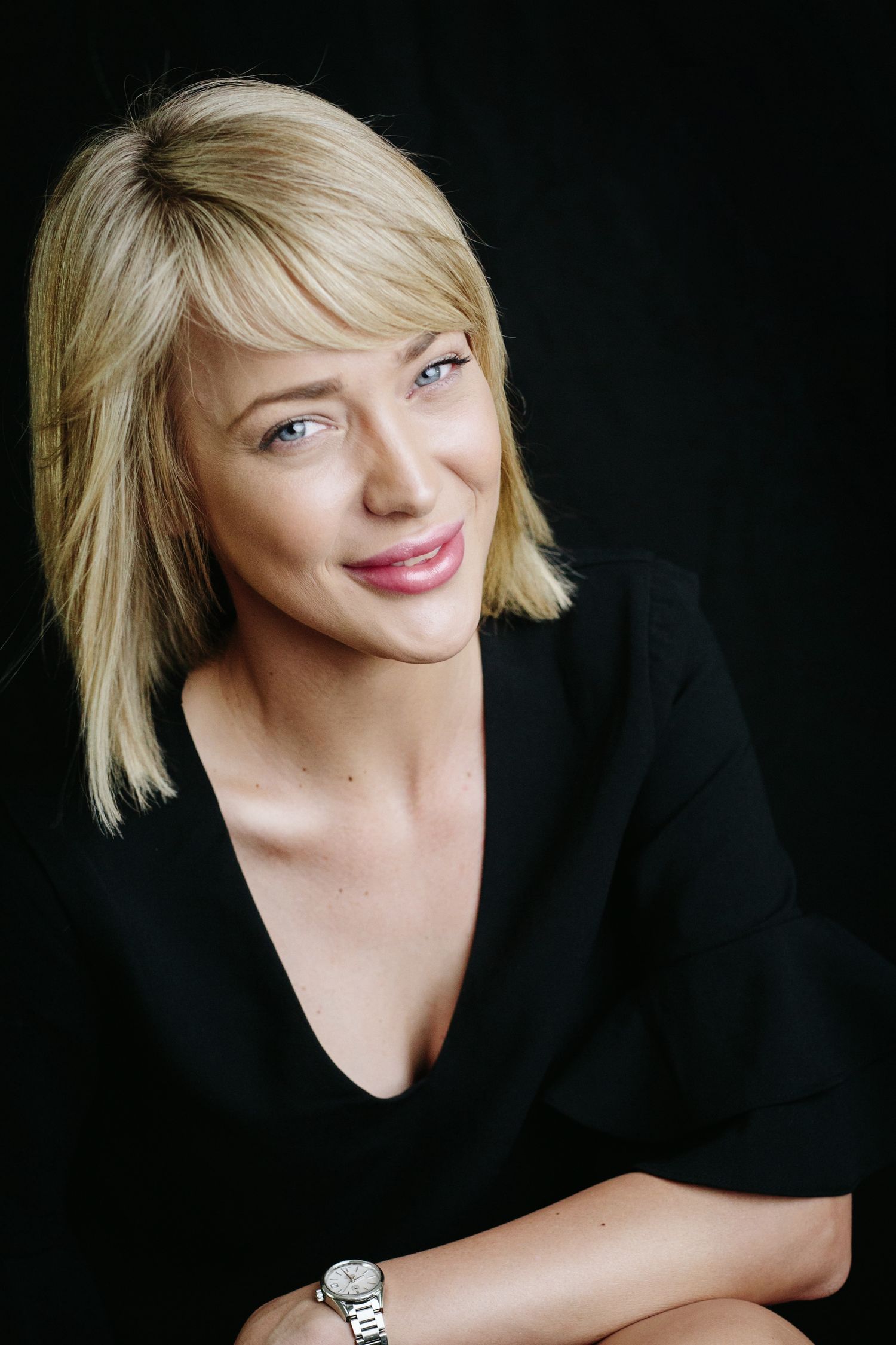 Smiling blonde woman with short hair wearing a black top and silver watch against dark background.