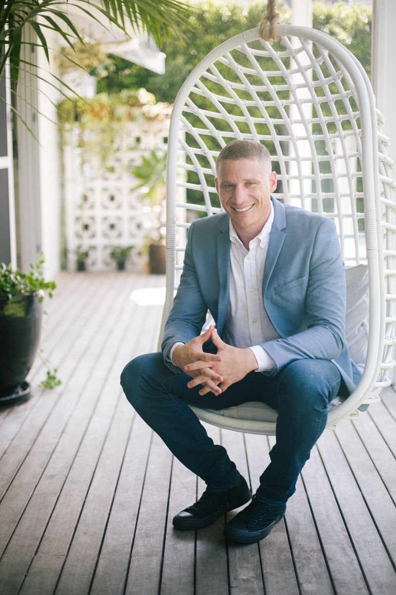 Smiling man in light blue blazer sitting in white wicker egg chair on wooden deck surrounded by greenery.