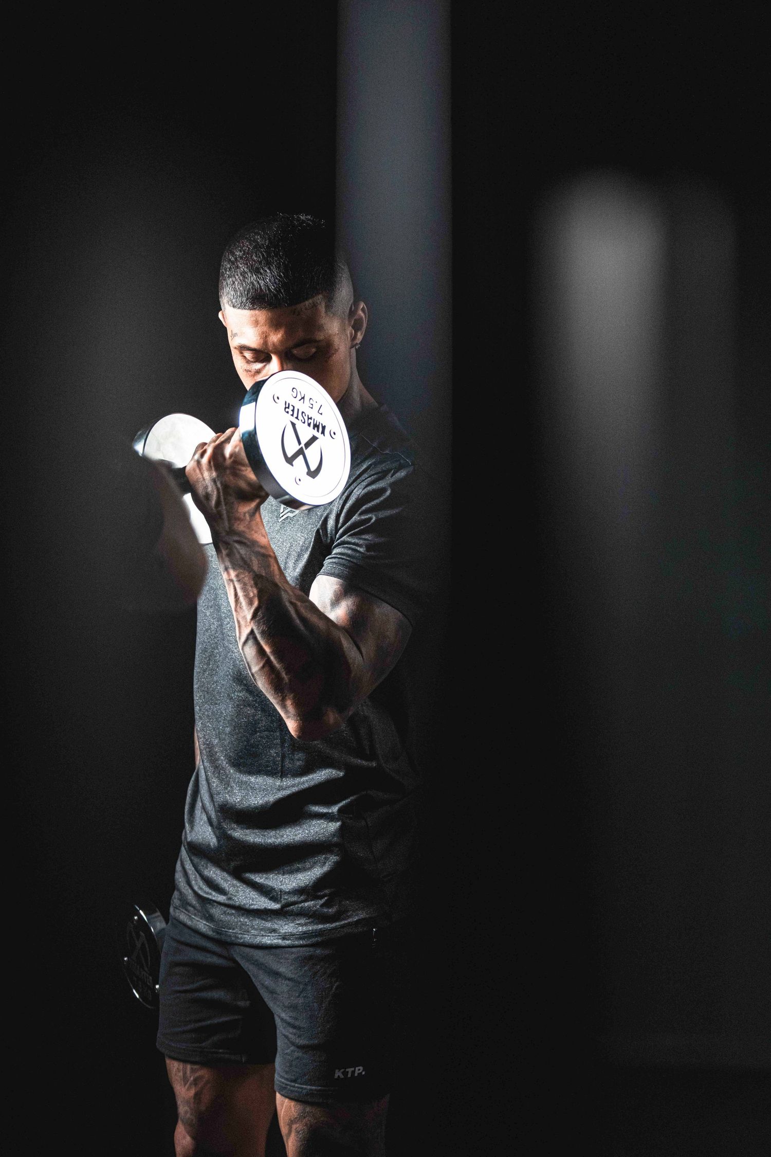 Muscular tattooed man in gray shirt curling a dumbbell in dramatic dark gym lighting.