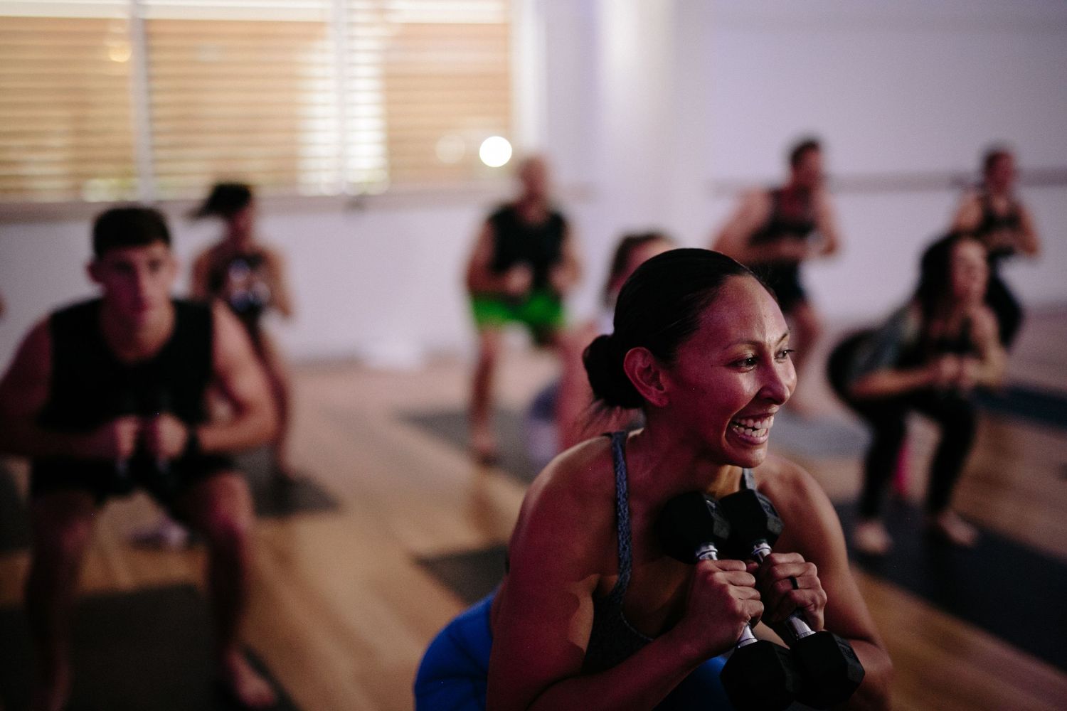 Group fitness class doing squats with weights in a dimly lit studio, focused woman in foreground grimacing with effort.
