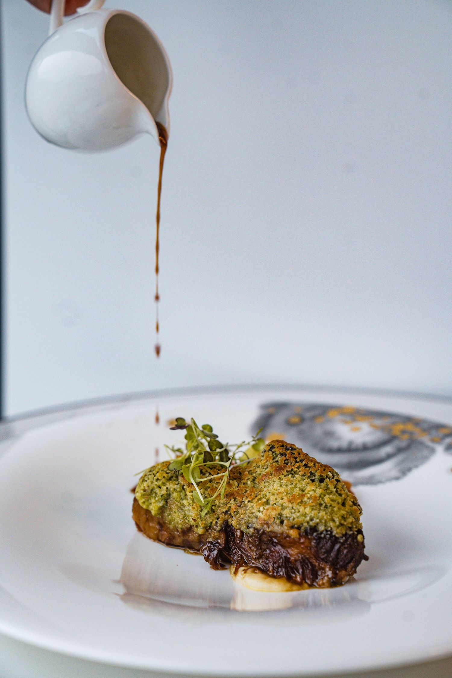 Rich brown sauce being poured over herb-crusted meat with garnishes on a pristine white plate.