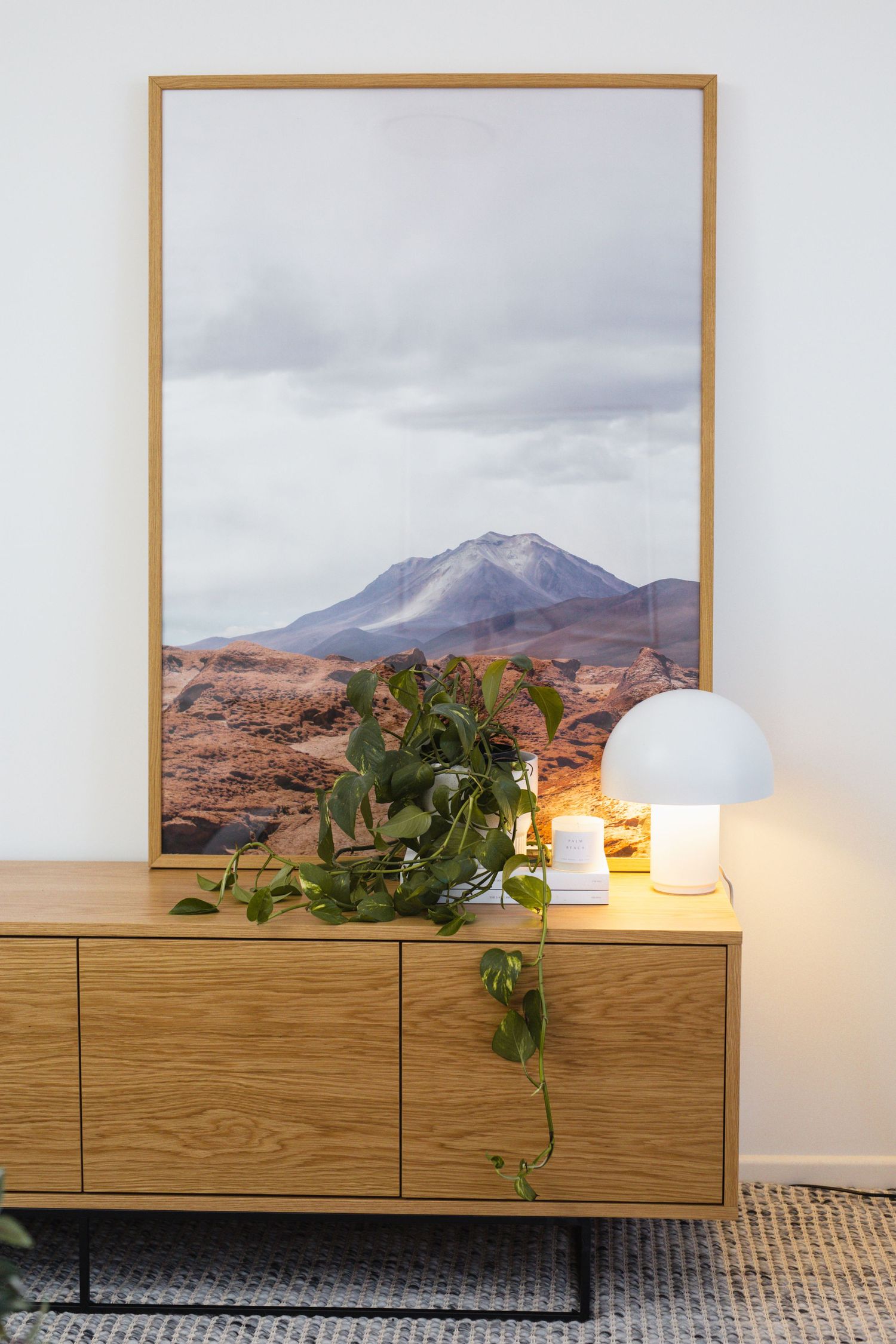 Modern oak sideboard with trailing pothos plant, glowing white mushroom lamp, and large framed mountain landscape artwork on white wall.