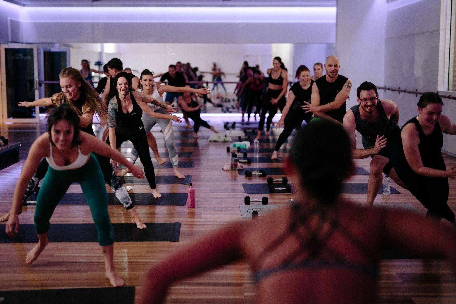 Group fitness class participants performing synchronized exercises in a modern gym studio with mirrors and dumbbells.