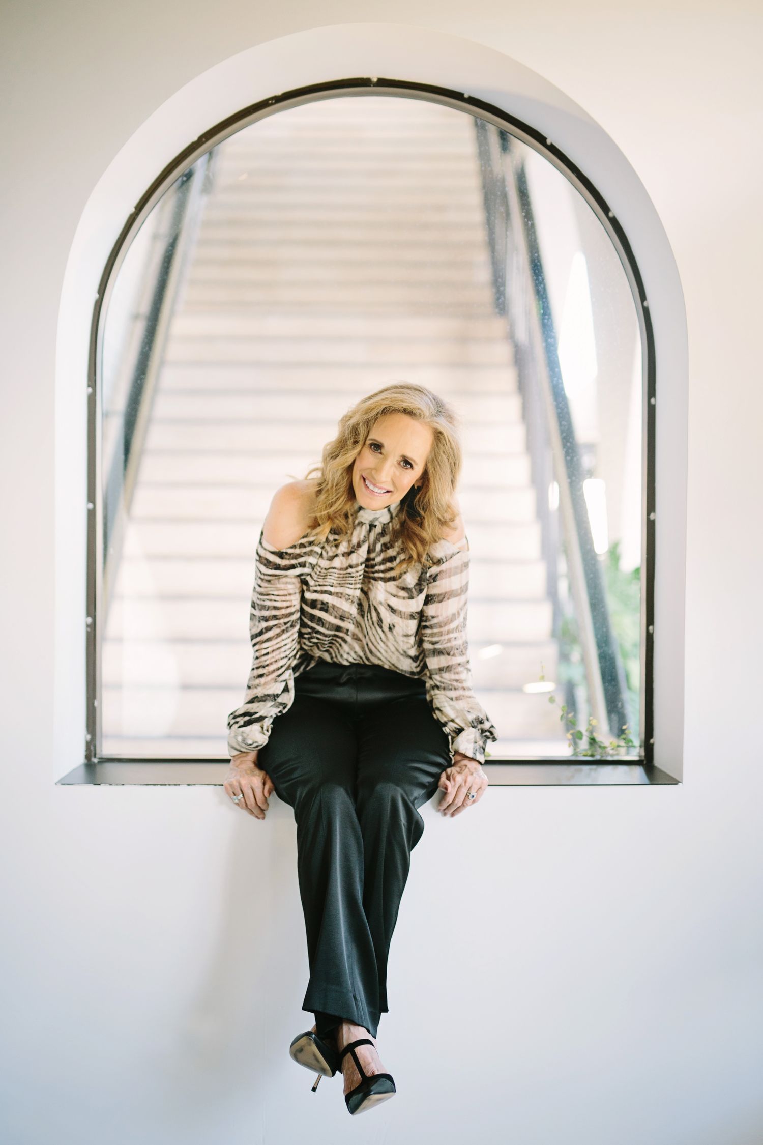 Smiling blonde woman sitting in arched window wearing tiger print off-shoulder top and black pants.