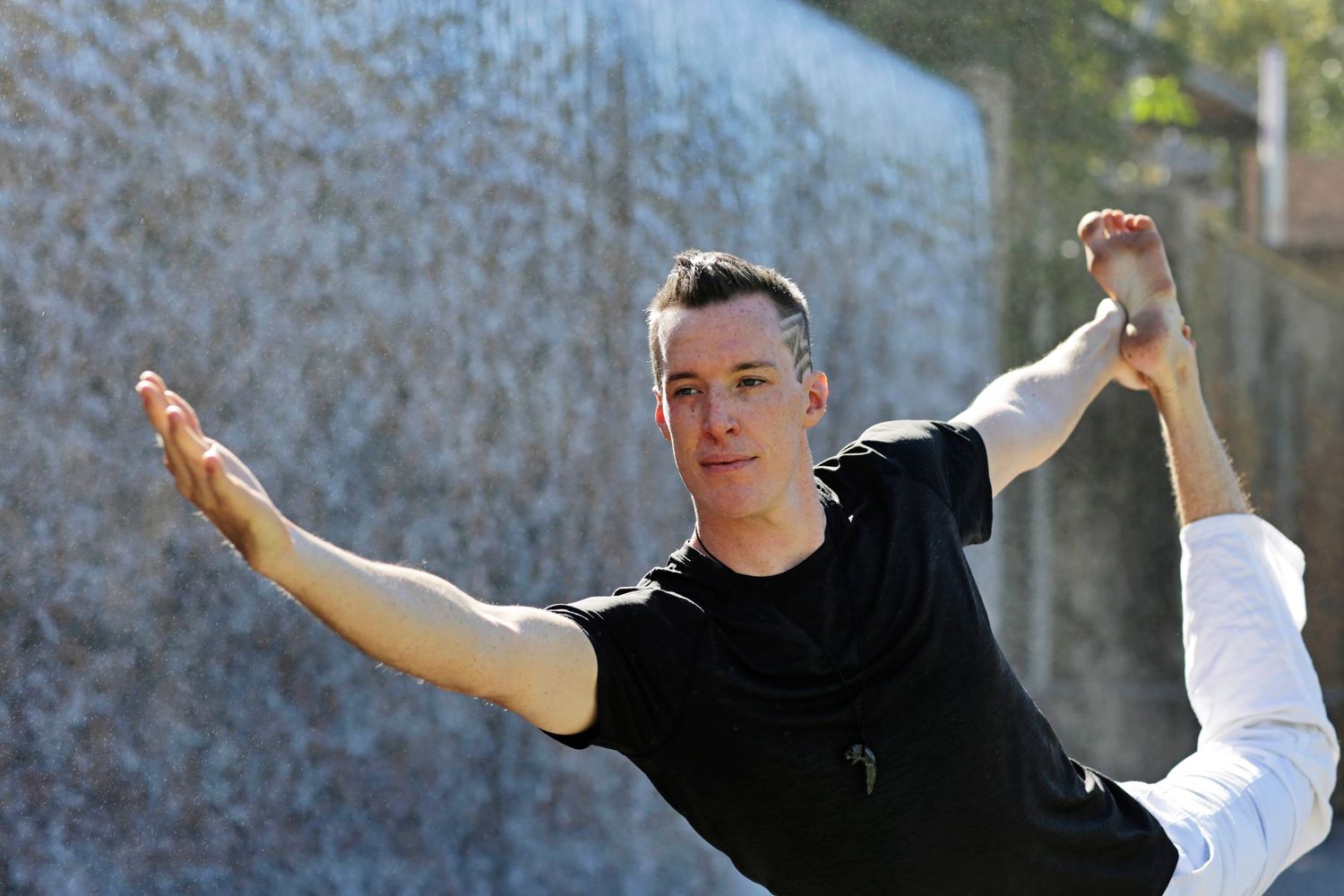 A young man in a black shirt performs a martial arts or dance pose outdoors near a stone wall.