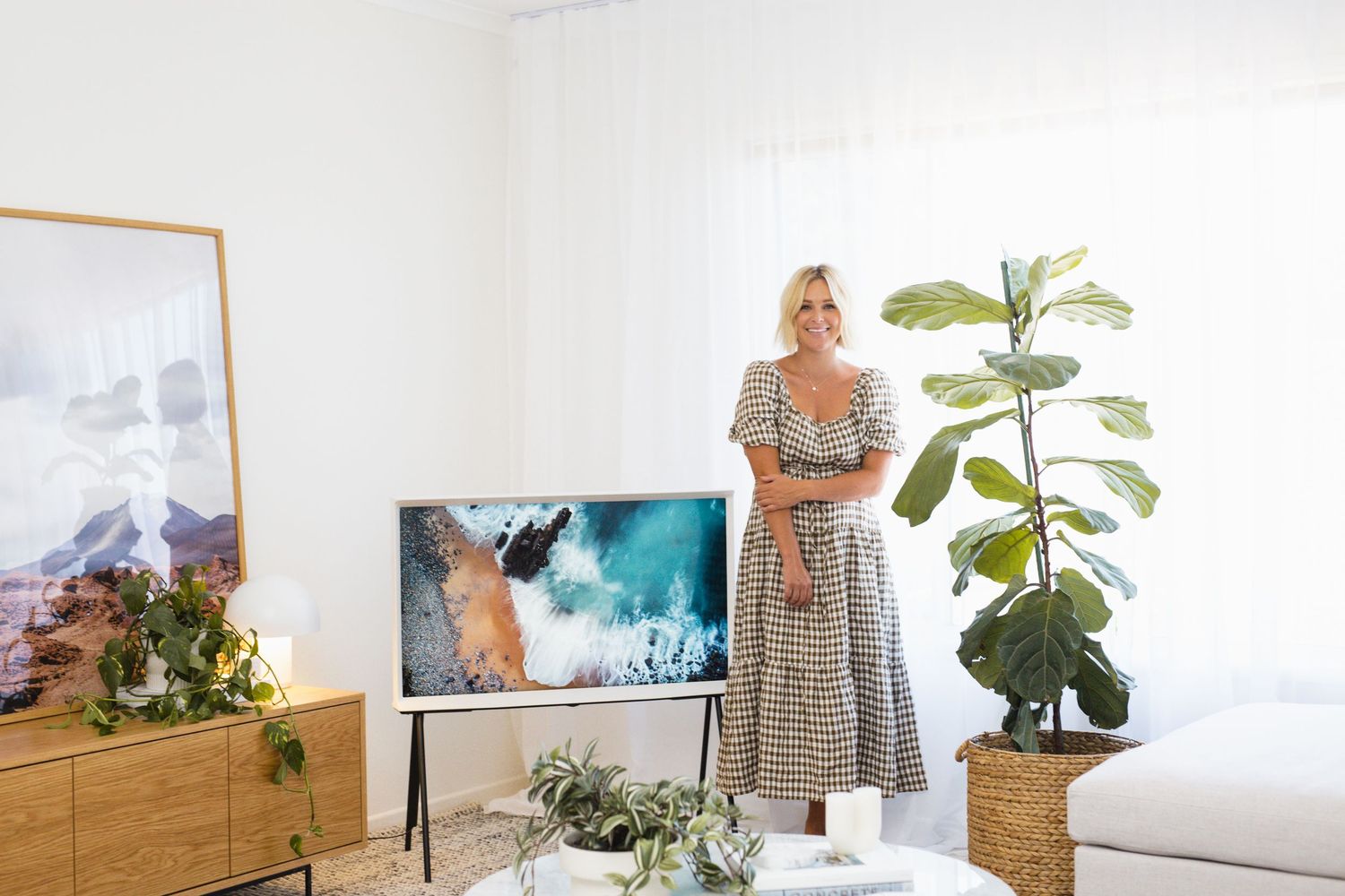 Woman in floral dress standing beside Samsung The Frame TV in bright modern living room with plants.