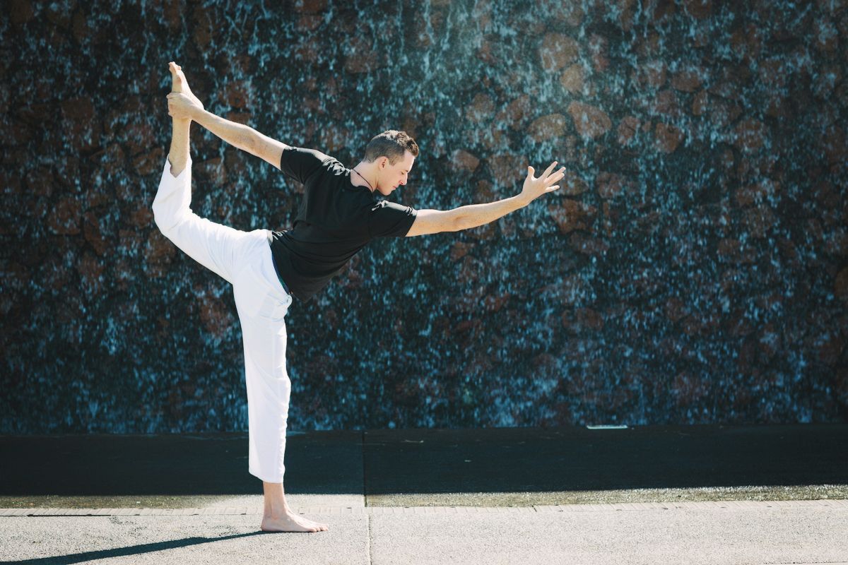 Person performing Lord of the Dance yoga pose outdoors in black top and white pants against textured wall.