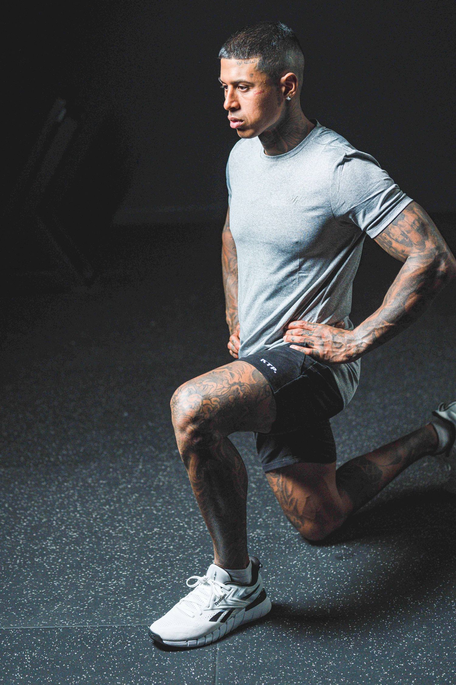 Tattooed male athlete performing a lunge exercise in a dark gym, wearing gray t-shirt and black shorts.