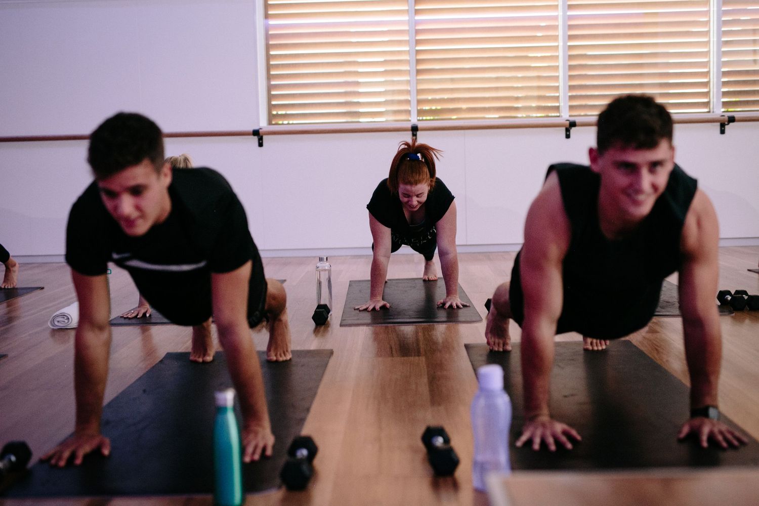 Three people doing push-ups on yoga mats in a gym with dumbbells and water bottles nearby.