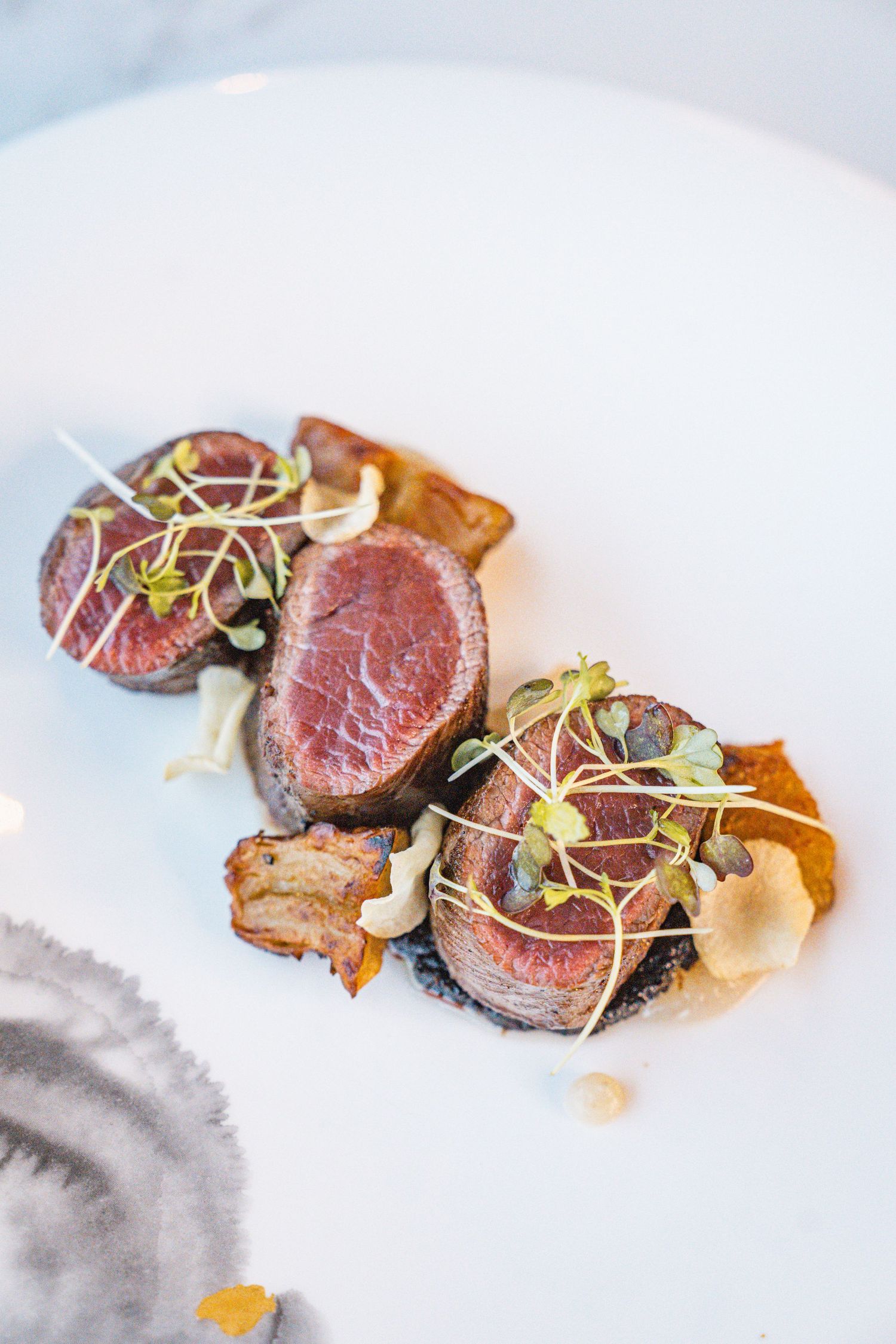 Sliced rare beef tenderloin garnished with microgreens and sauce on white plates shown from multiple angles.
