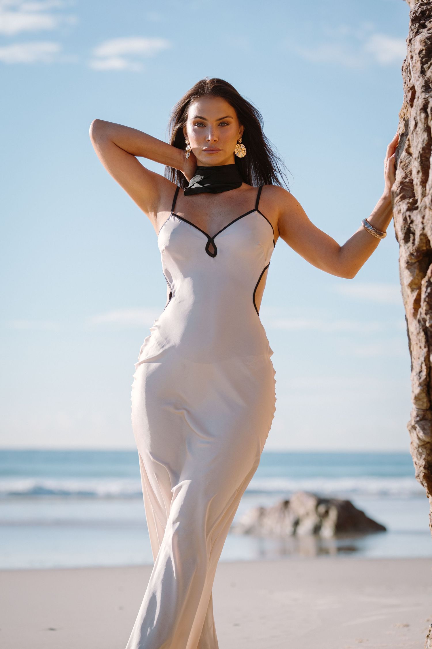 Elegant white evening gown with choker detail photographed against beach backdrop with ocean and rocks.