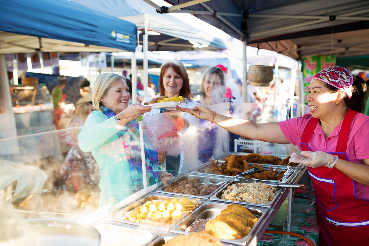 Customers selecting food from steam trays at an outdoor market food stall with a blue canopy overhead.