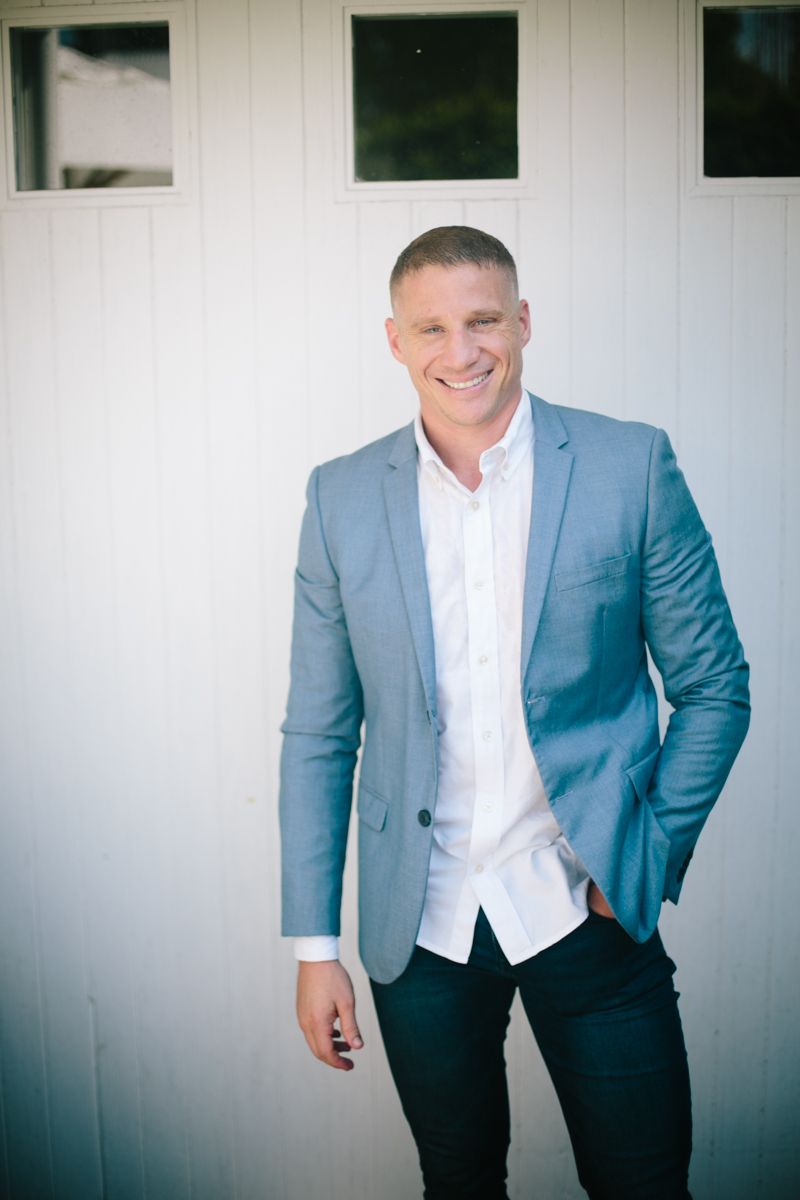 Smiling muscular man in light blue blazer and white shirt posing confidently against white barn door.