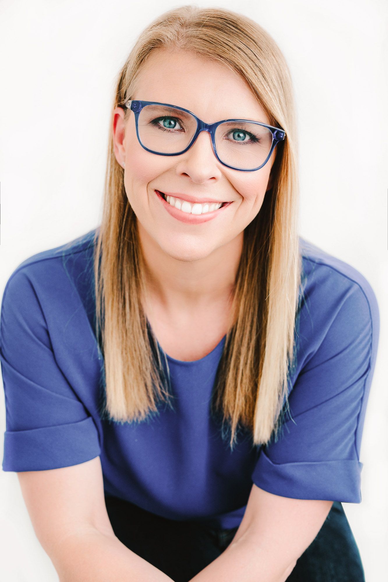 Professional headshot of person in blue top and glasses smiling warmly against white background.