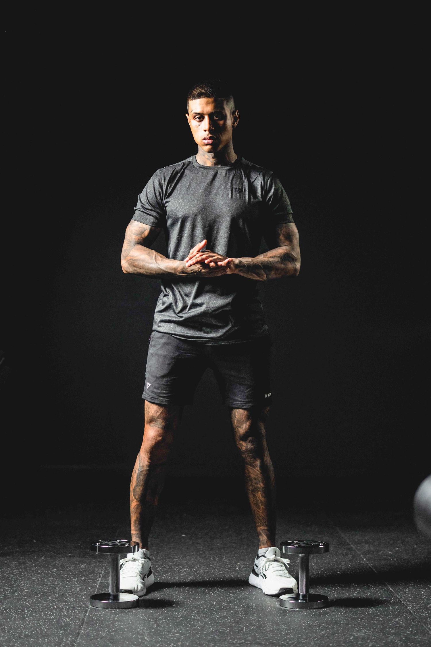 Muscular man in gray athletic wear stands confidently in gym with dumbbells, dark background.